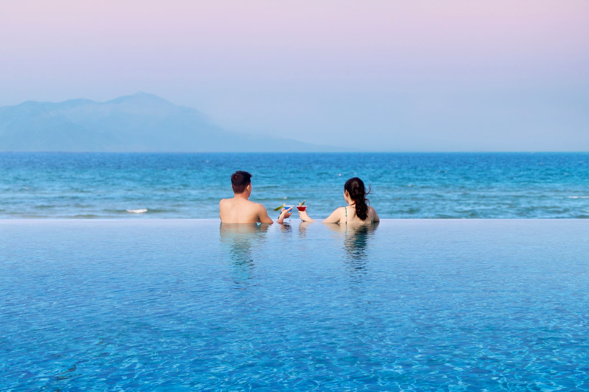 Relaxing couple sips cocktails in infinity pool with ocean and mountains.