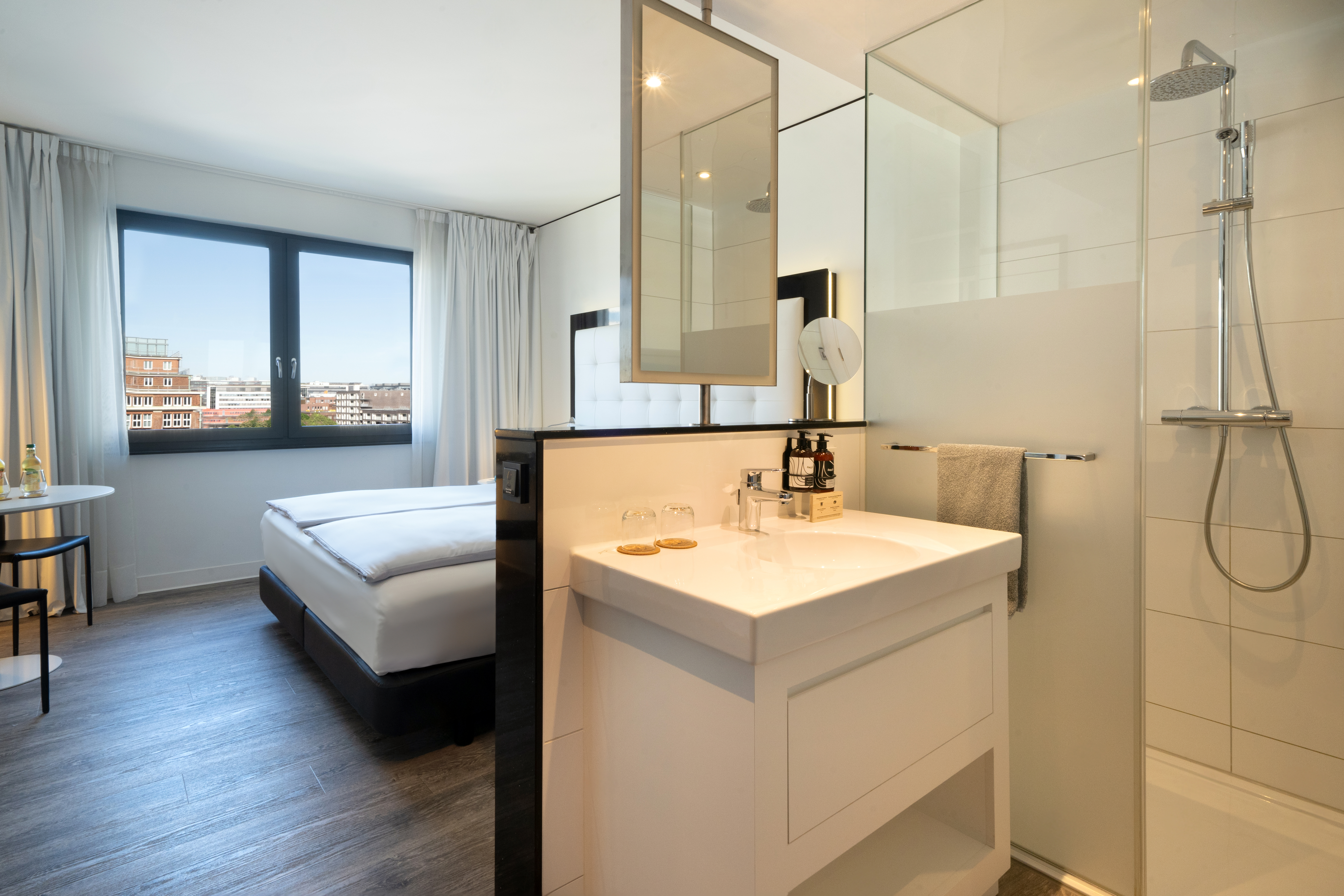 Modern hotel room with city view, bed, and open bathroom with sink and shower.