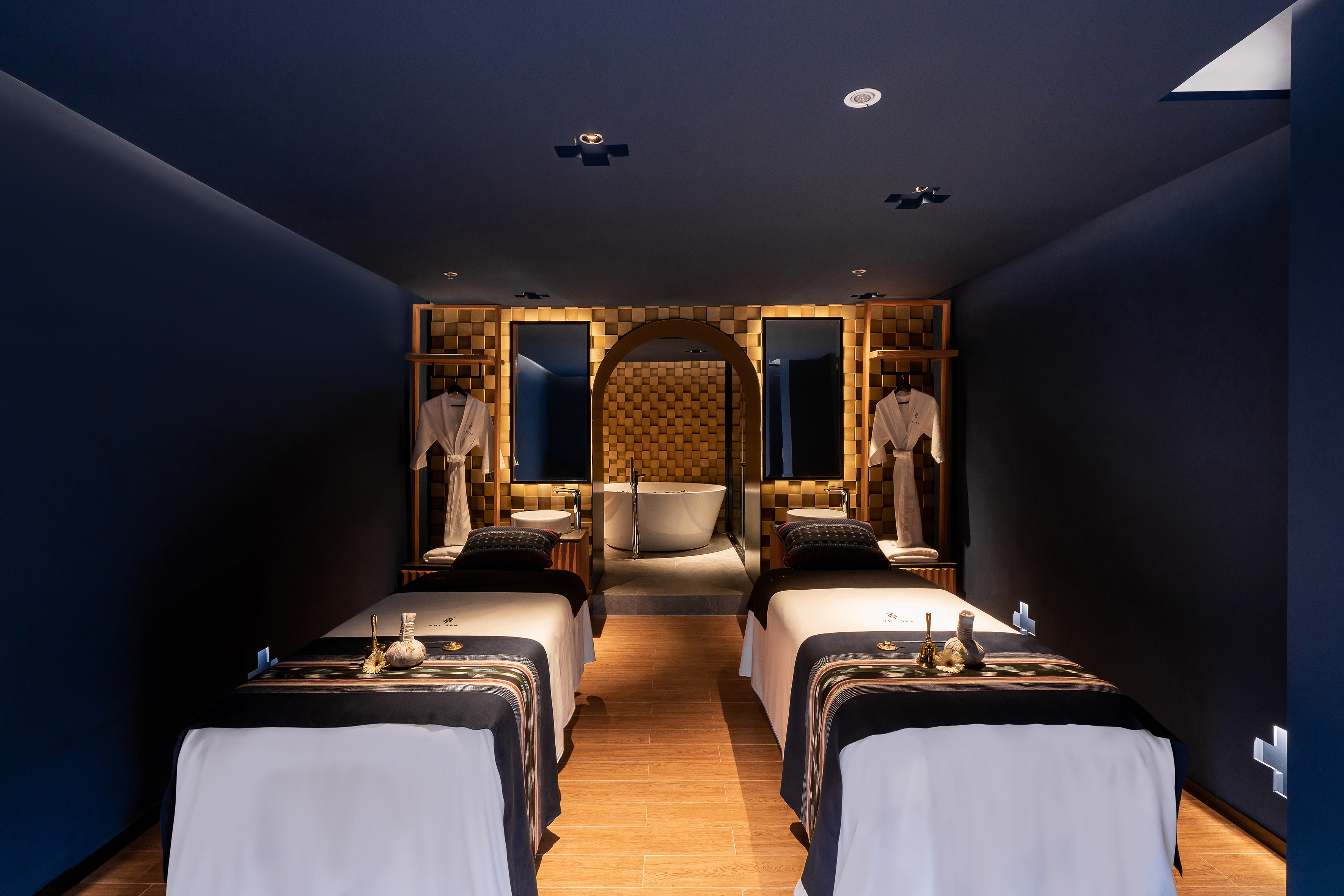 Luxury spa room with two massage beds, dark blue decor, and bathtub.