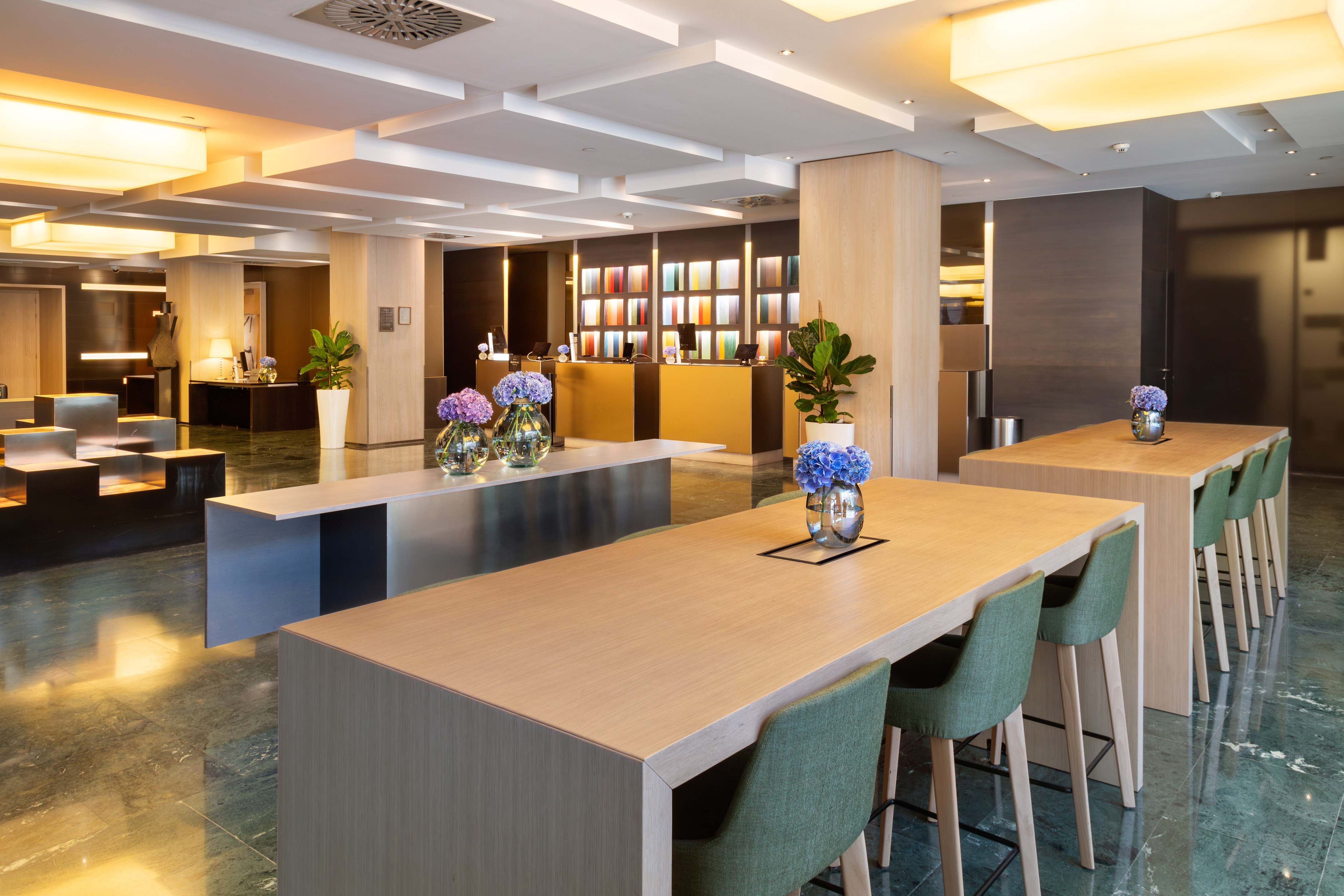 Modern hotel lobby, long tables, green stools, gold reception, and marble floor.
