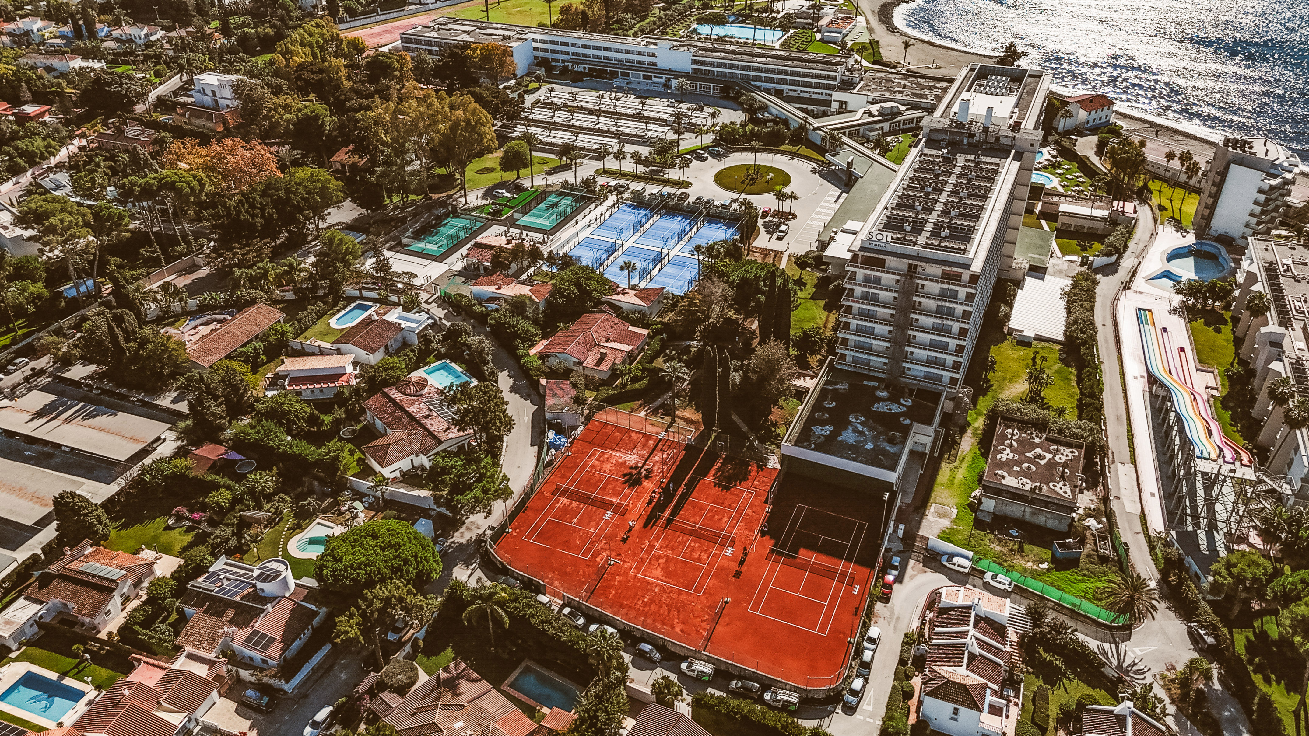 Aerial: Coastal resort with hotels, red tennis courts, blue paddle courts, and beach.