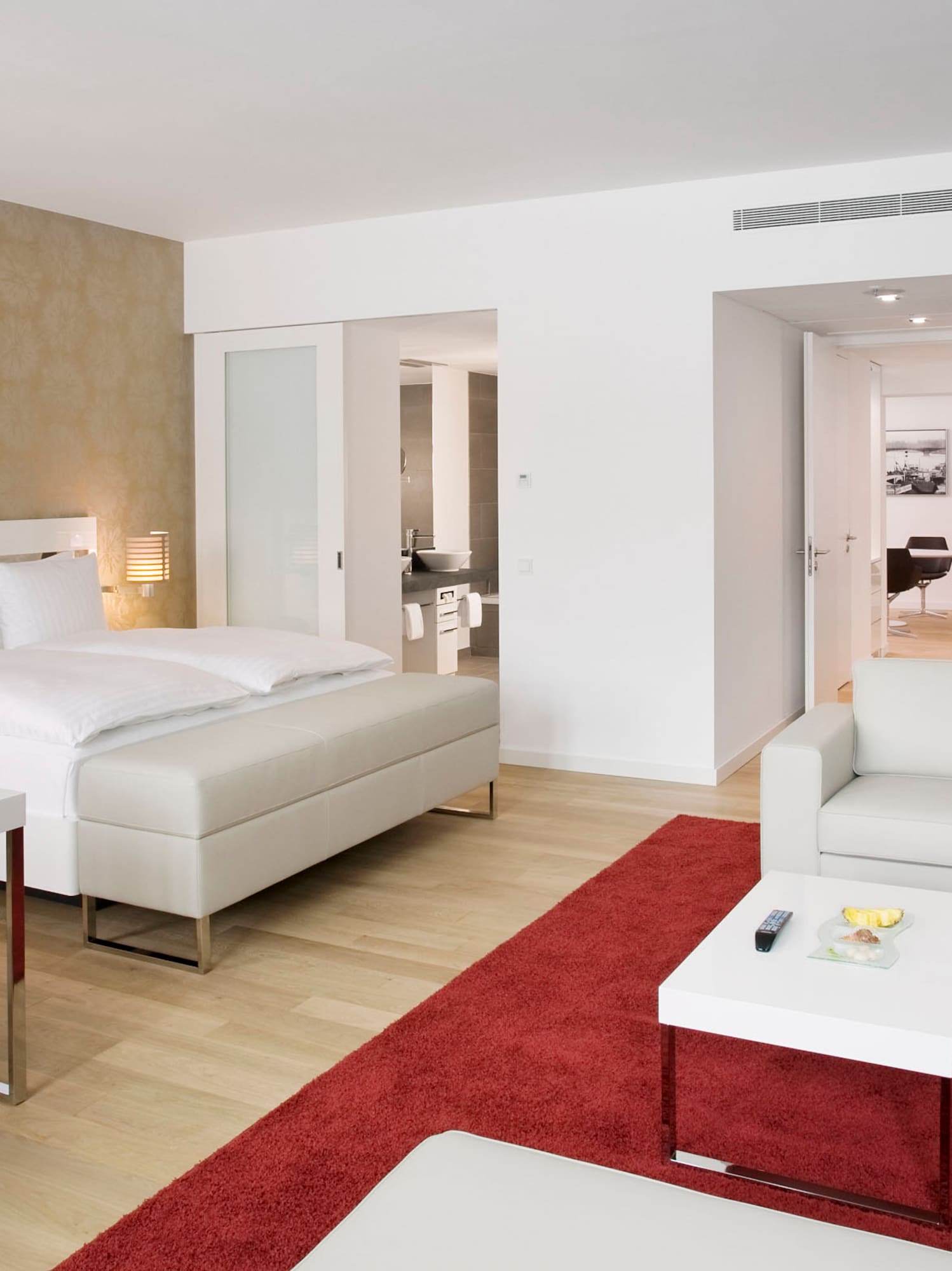 Modern hotel suite with white bed, red rug, seating area, and desk.