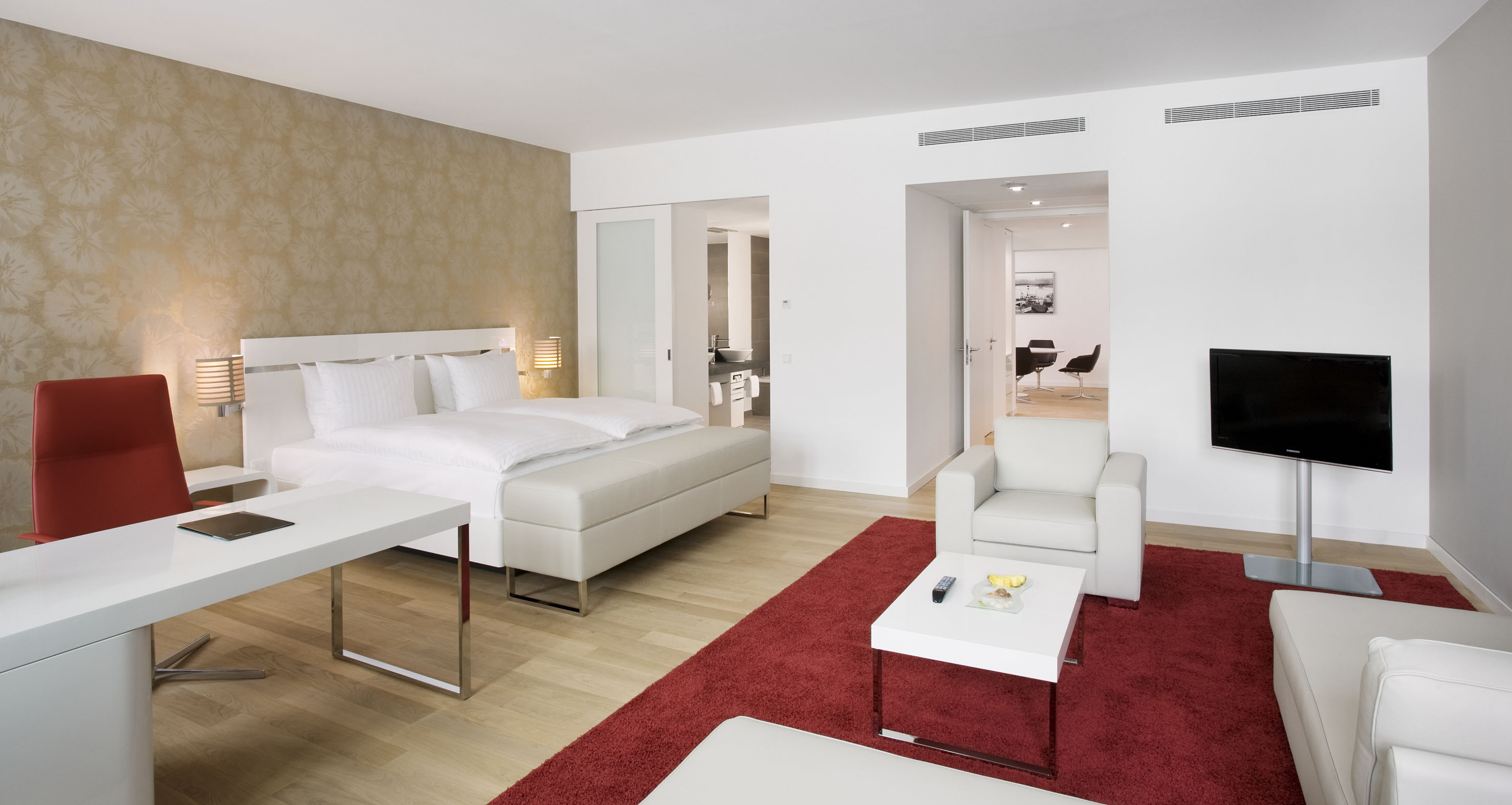 Modern hotel suite with white bed, red rug, seating area, and desk.
