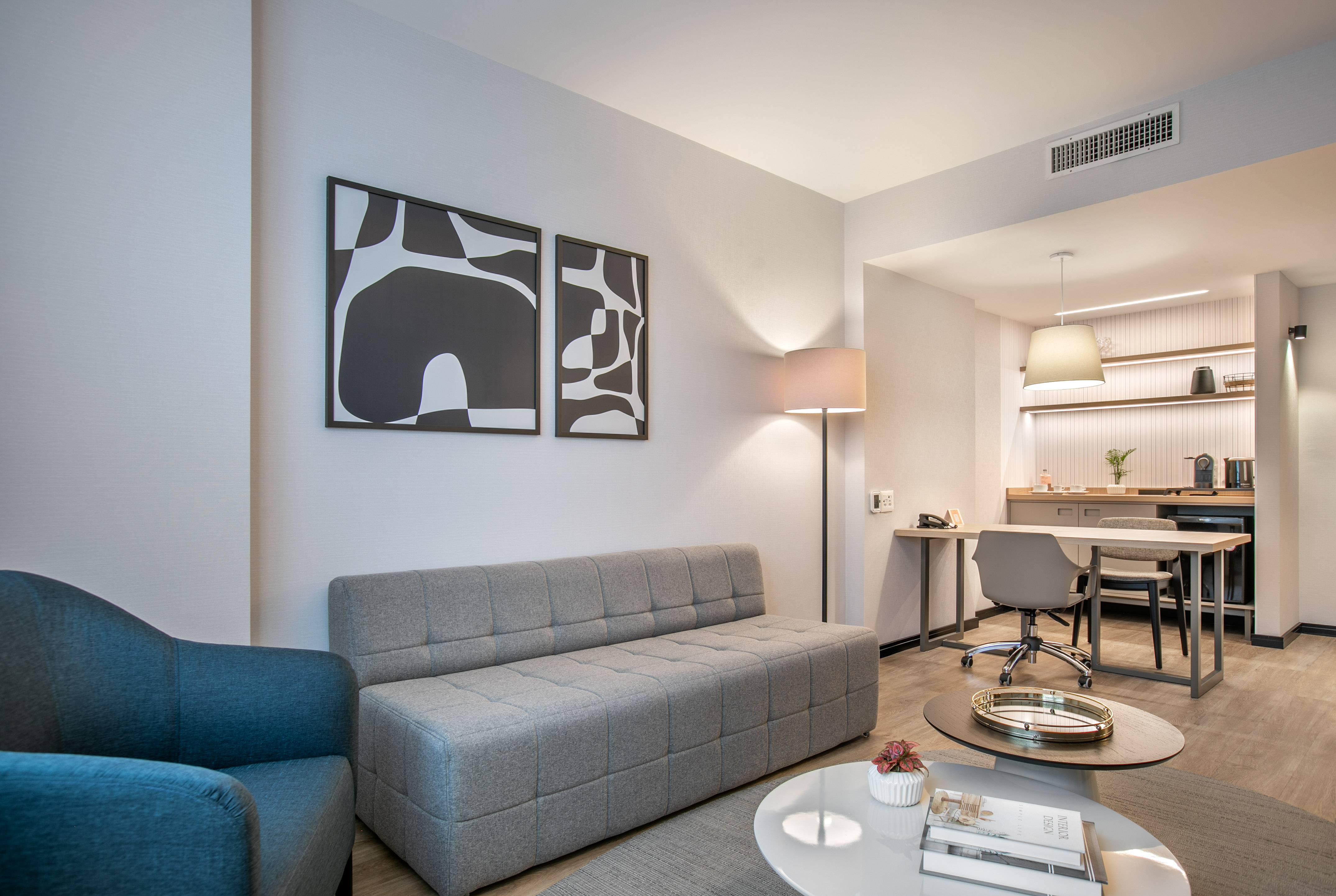 Modern hotel suite living area with grey sofa, abstract art, desk, and kitchenette.