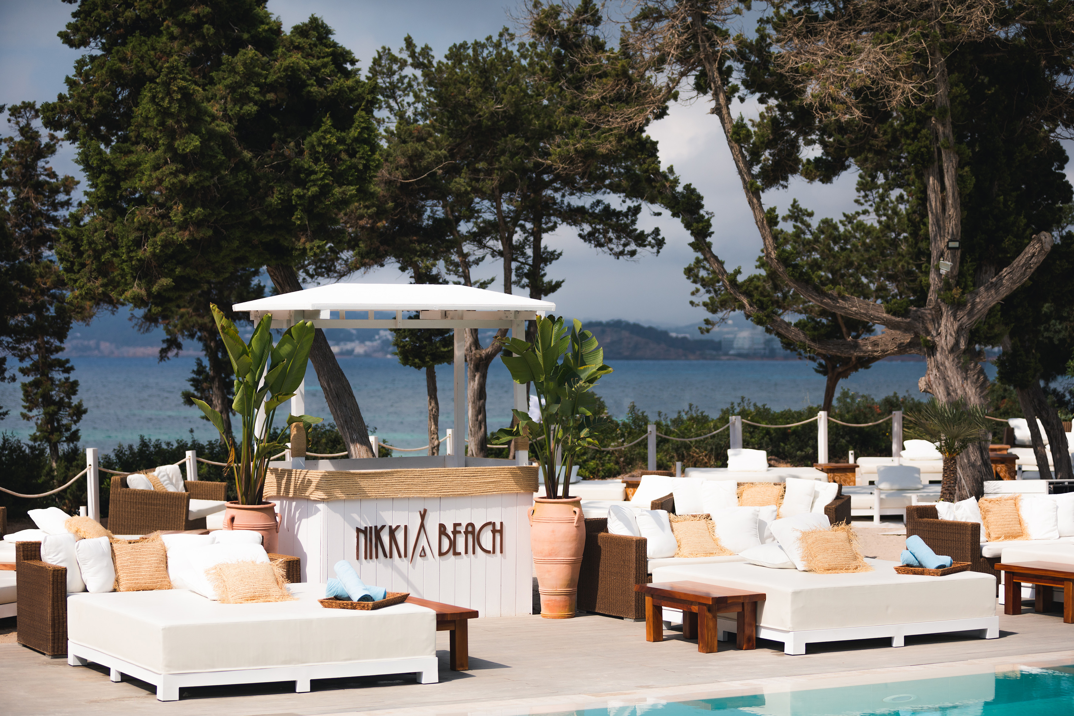 Nikki Beach bar, white daybeds, and ocean view by lush cypress trees.