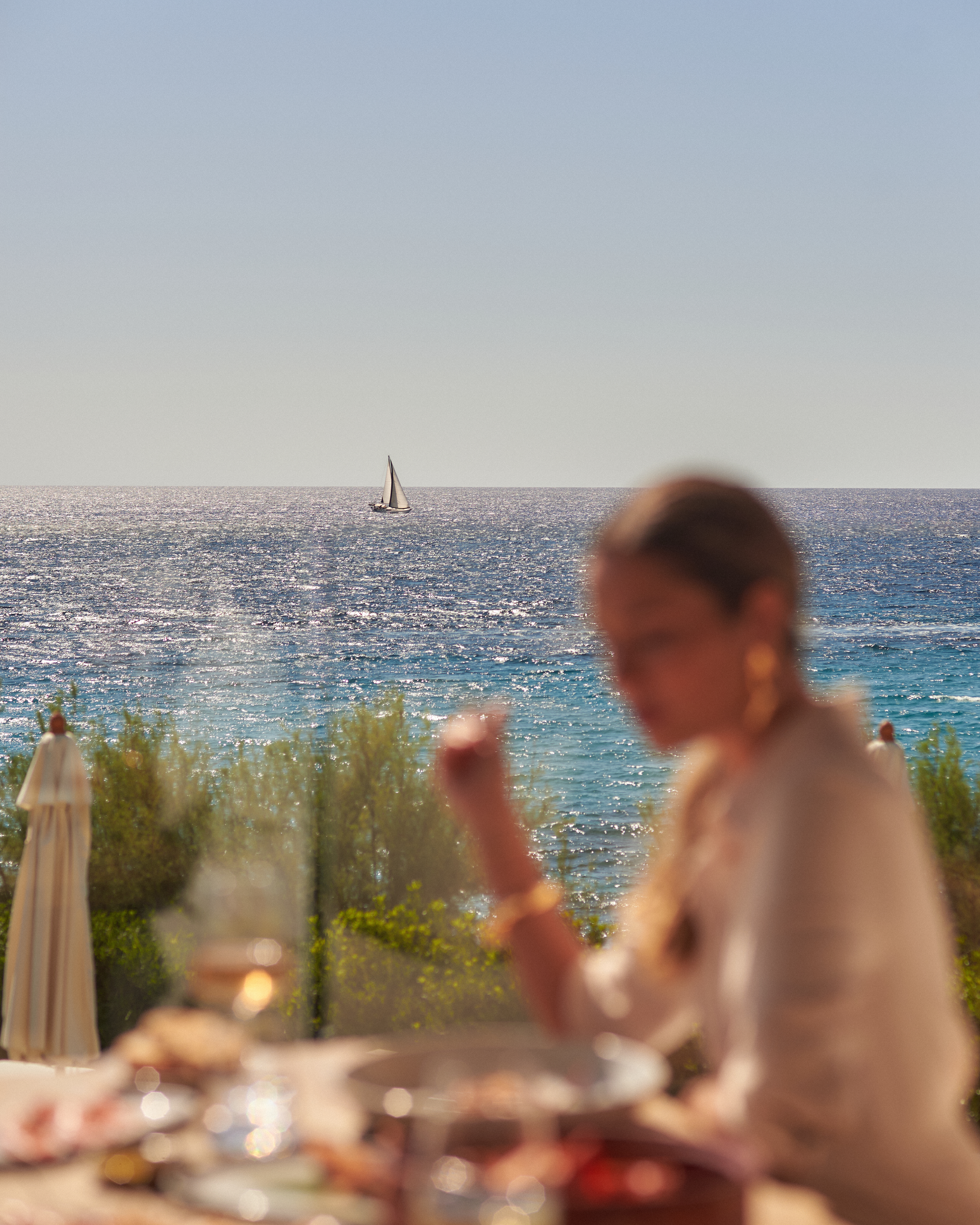 Sailboat on sparkling sea beyond blurred outdoor dining table.