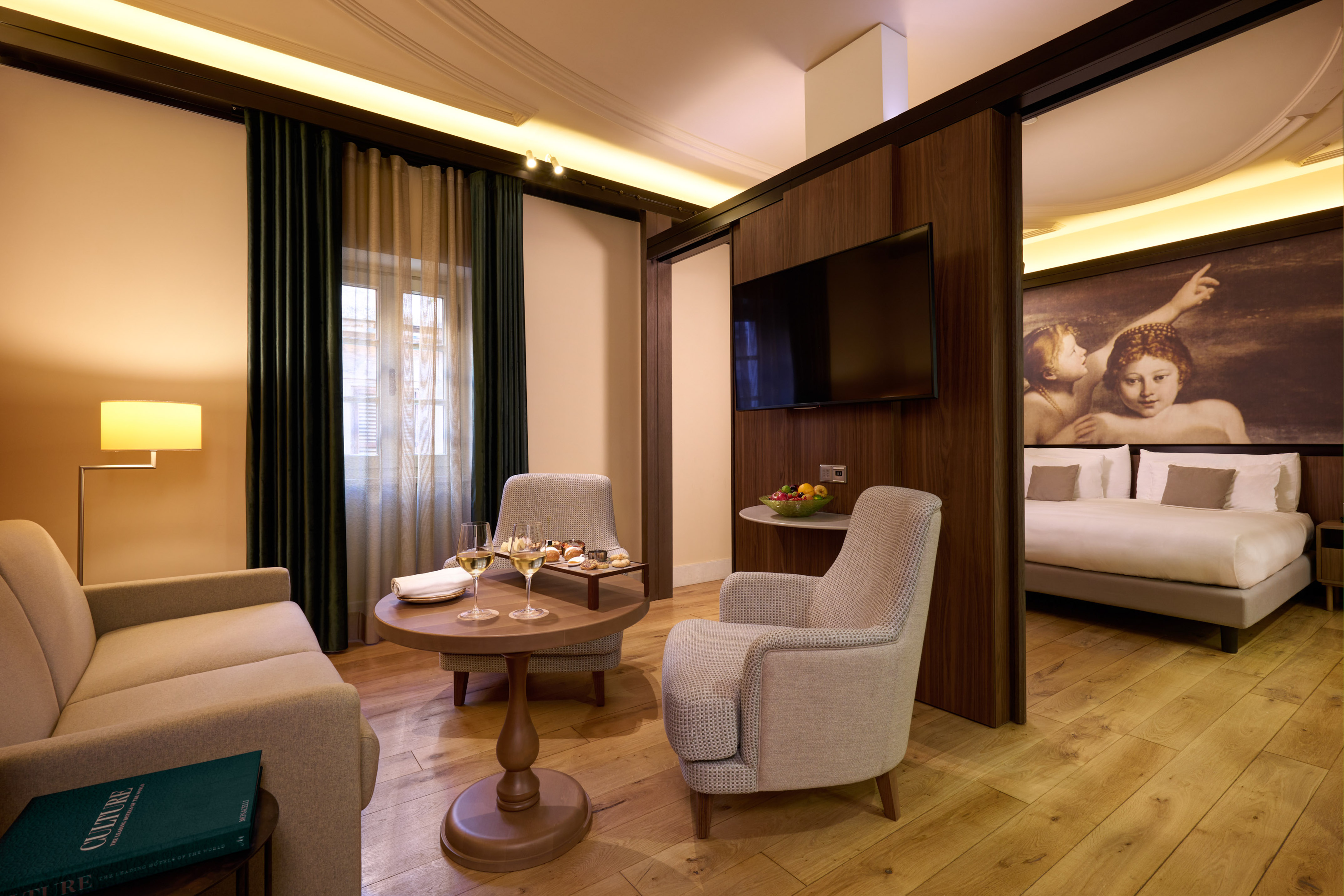 Modern hotel suite with living area, bedroom, wood floors, wine, and TV.