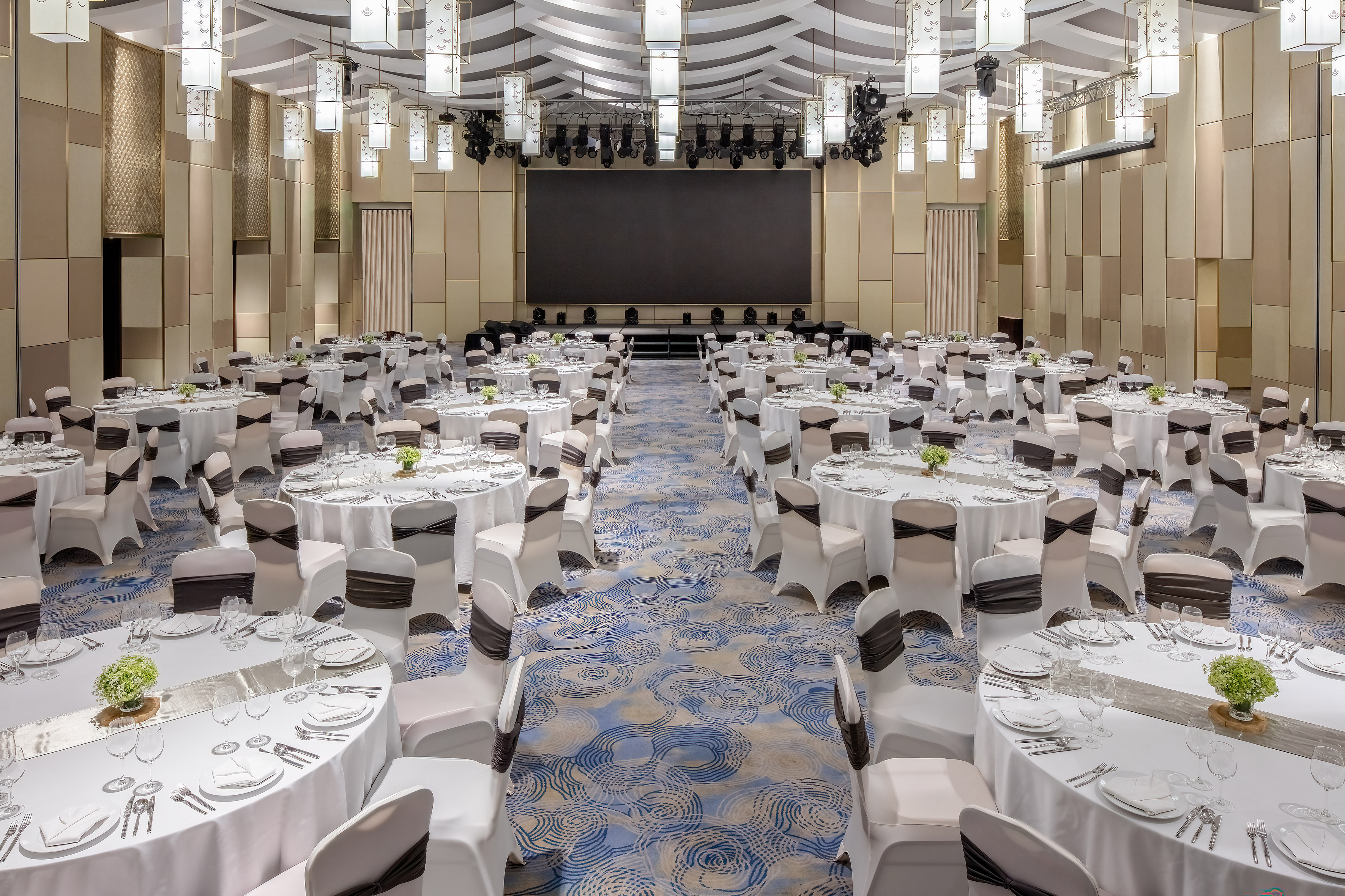 Elegant banquet hall with round tables, white chairs, stage, and modern lighting.