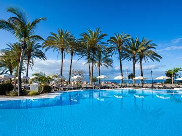 Tropical resort pool with palm trees, loungers, umbrellas, and ocean view.