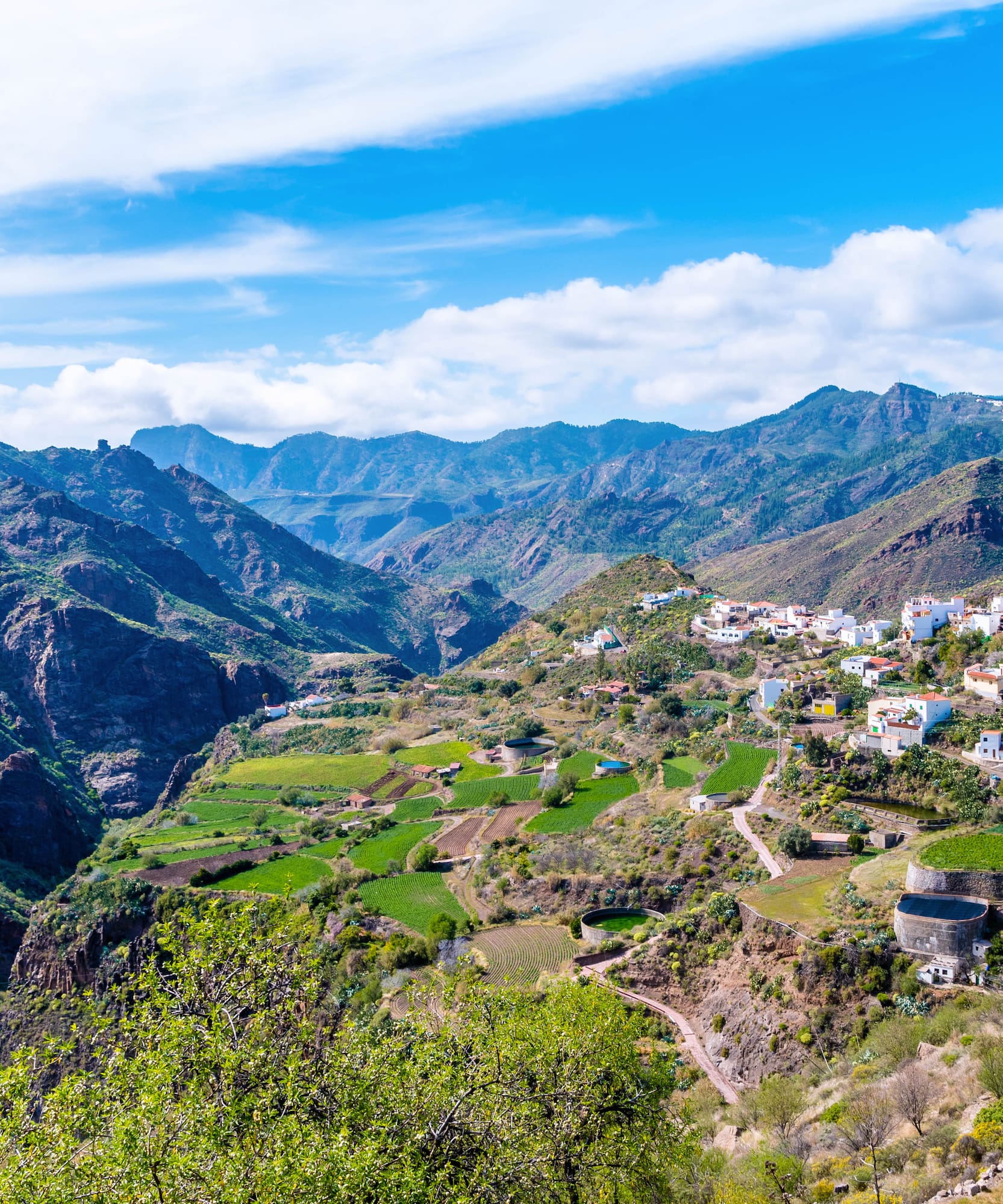 Pueblo blanco en valle montañoso, campos verdes y cielo azul.