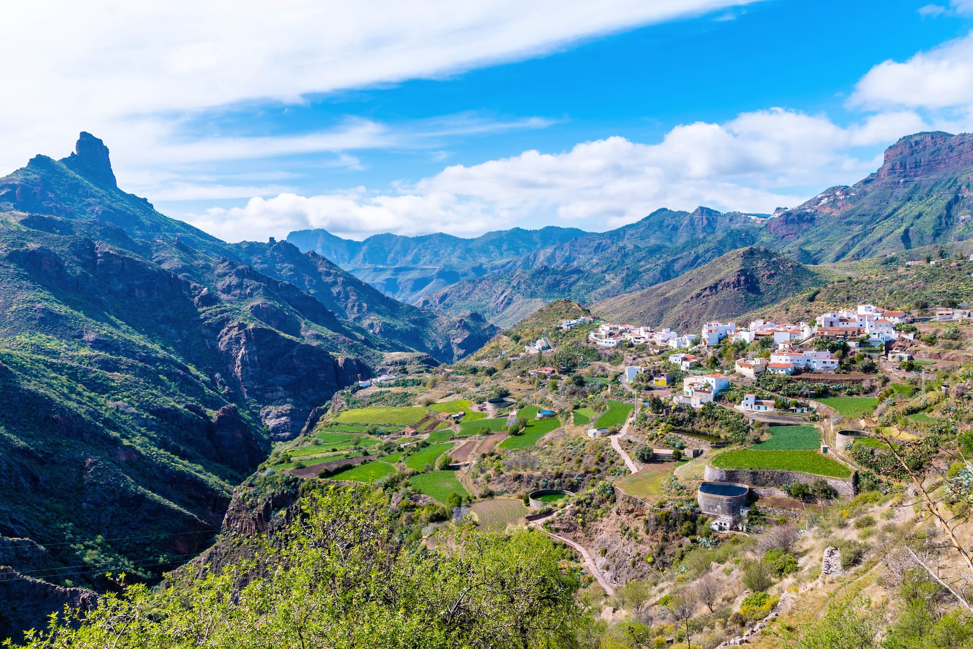 Pueblo blanco en valle montañoso, campos verdes y cielo azul.