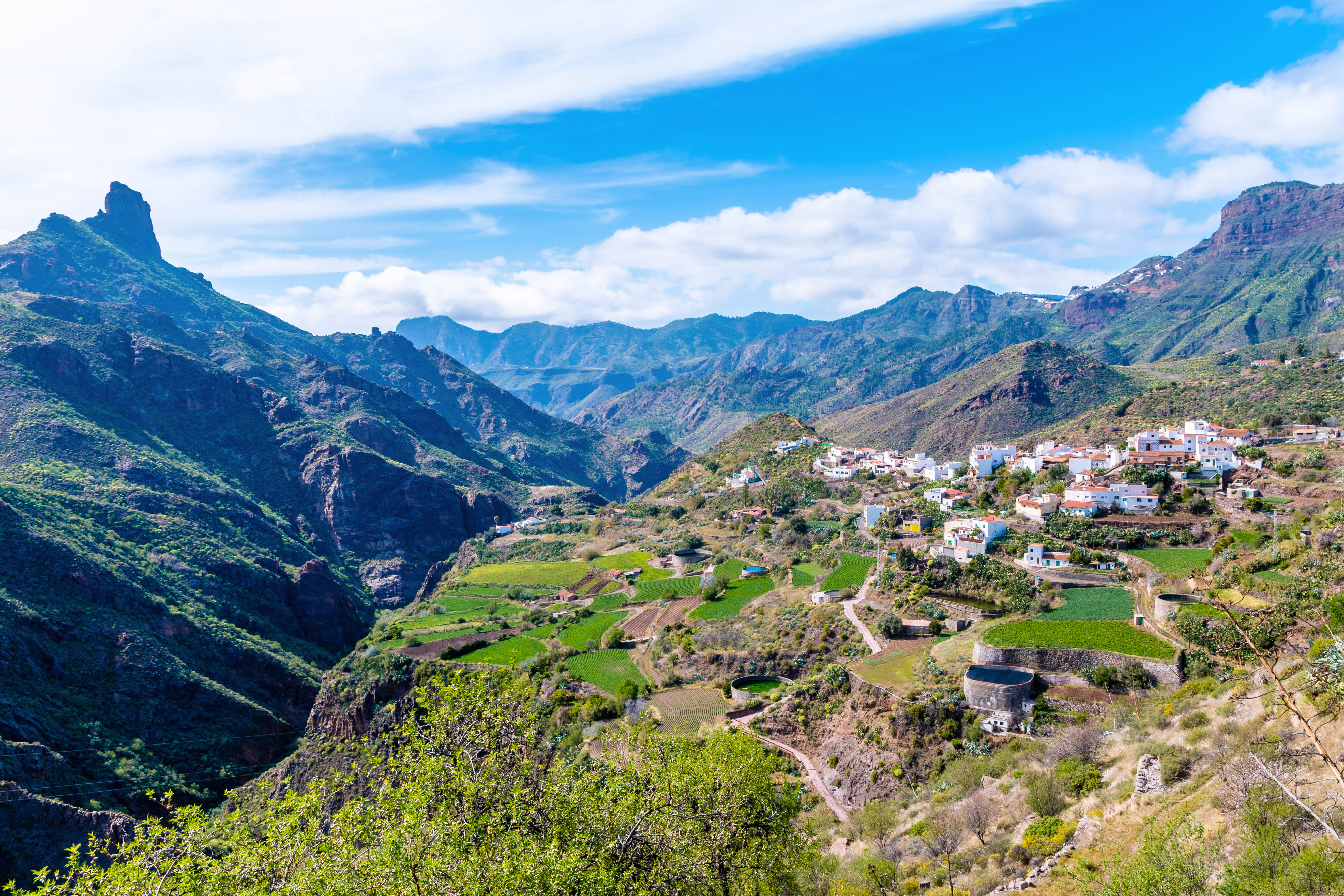 Pueblo blanco en valle montañoso, campos verdes y cielo azul.