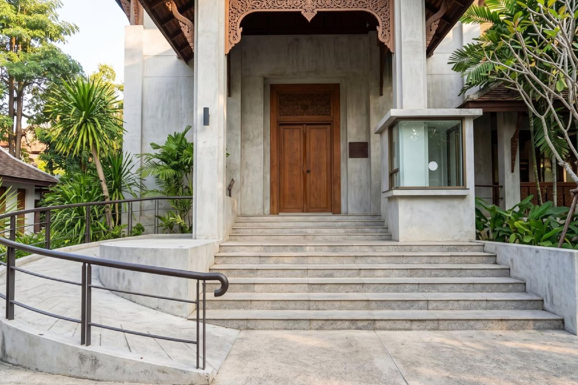 Entrada edificio con puertas de madera tallada, escaleras, rampa y jardín tropical.