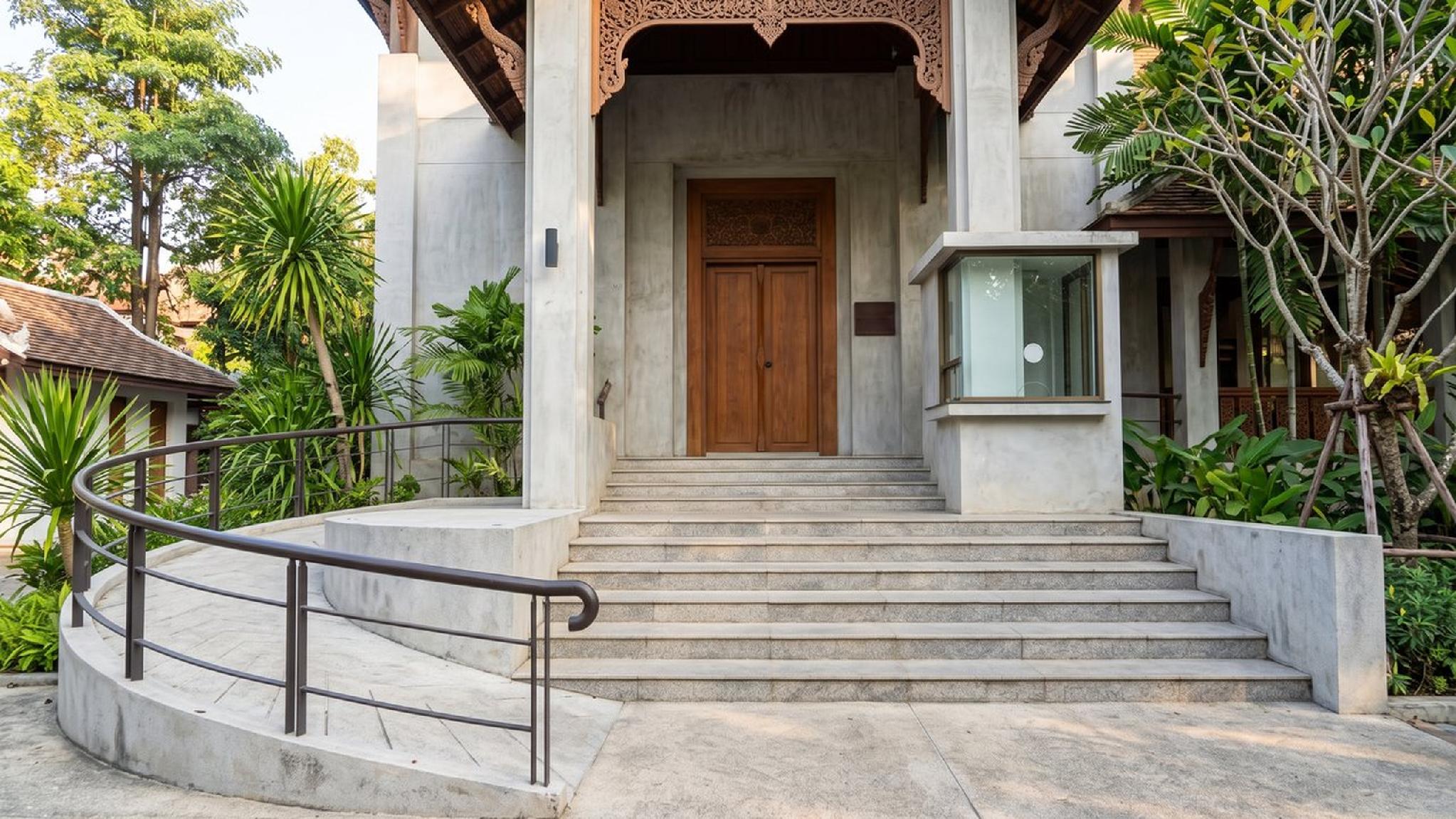Entrada edificio con puertas de madera tallada, escaleras, rampa y jardín tropical.