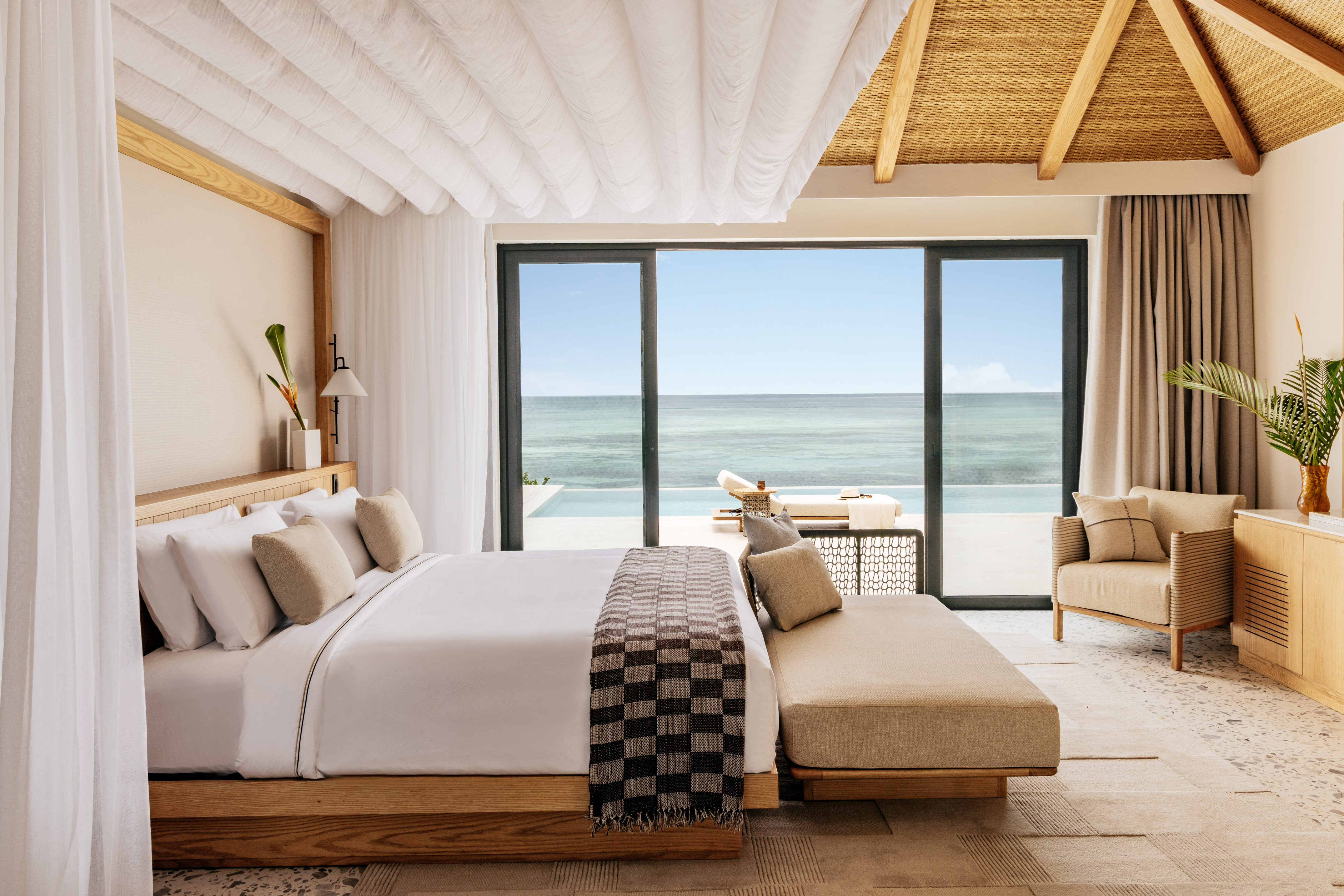 Luxury beachfront bedroom with canopy bed, infinity pool, and thatched ceiling.