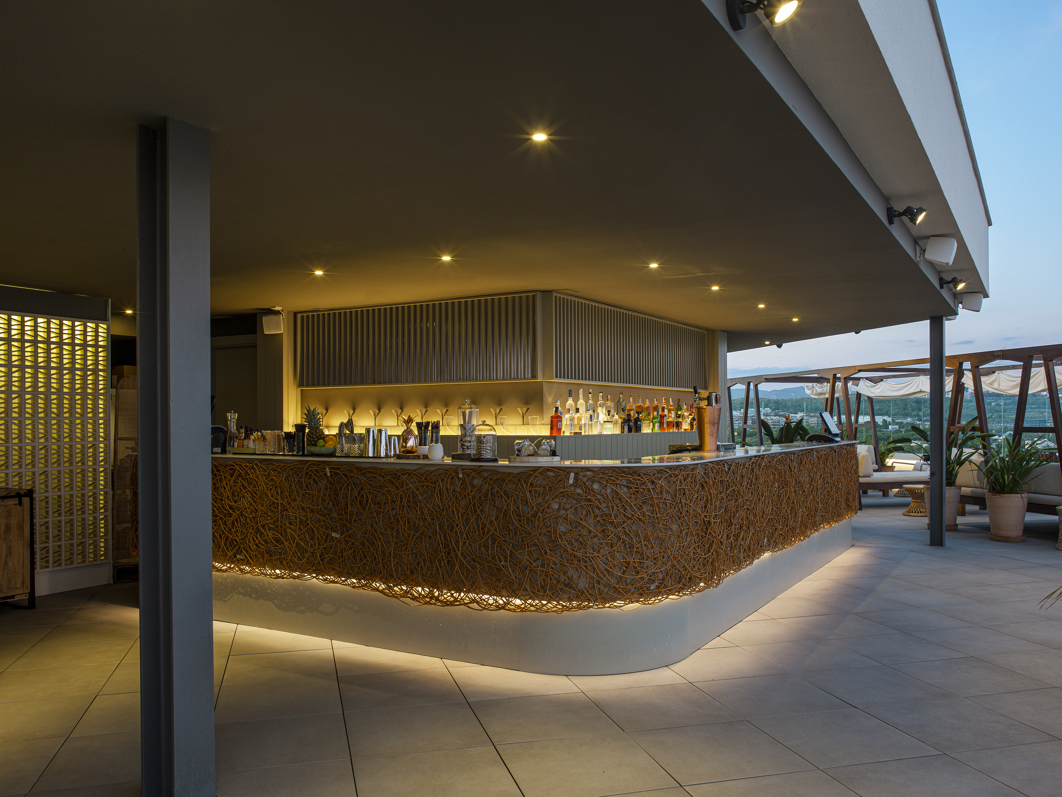 Rooftop bar with woven counter, under-lighting, and outdoor lounge at dusk.