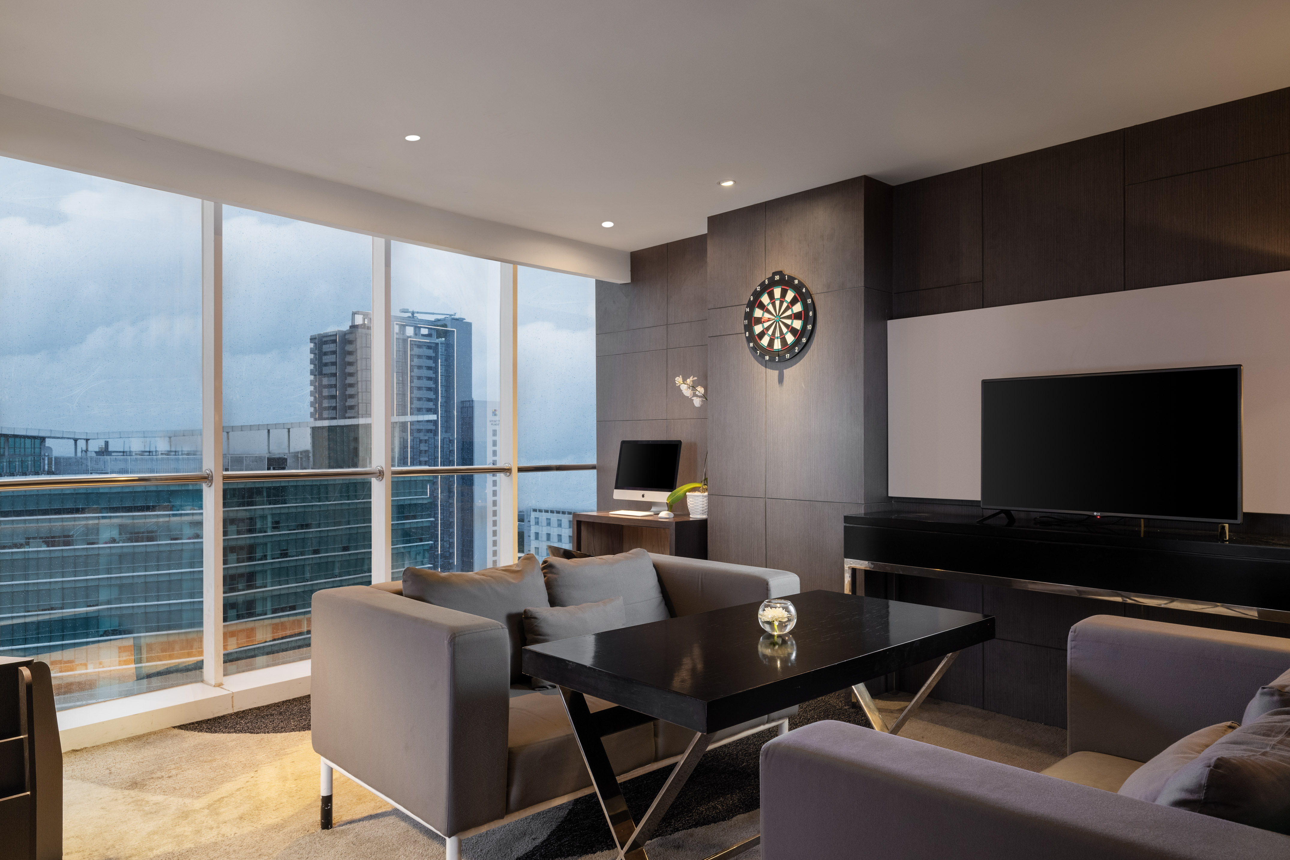 Modern living room with city view, grey sofas, dartboard, and desk.
