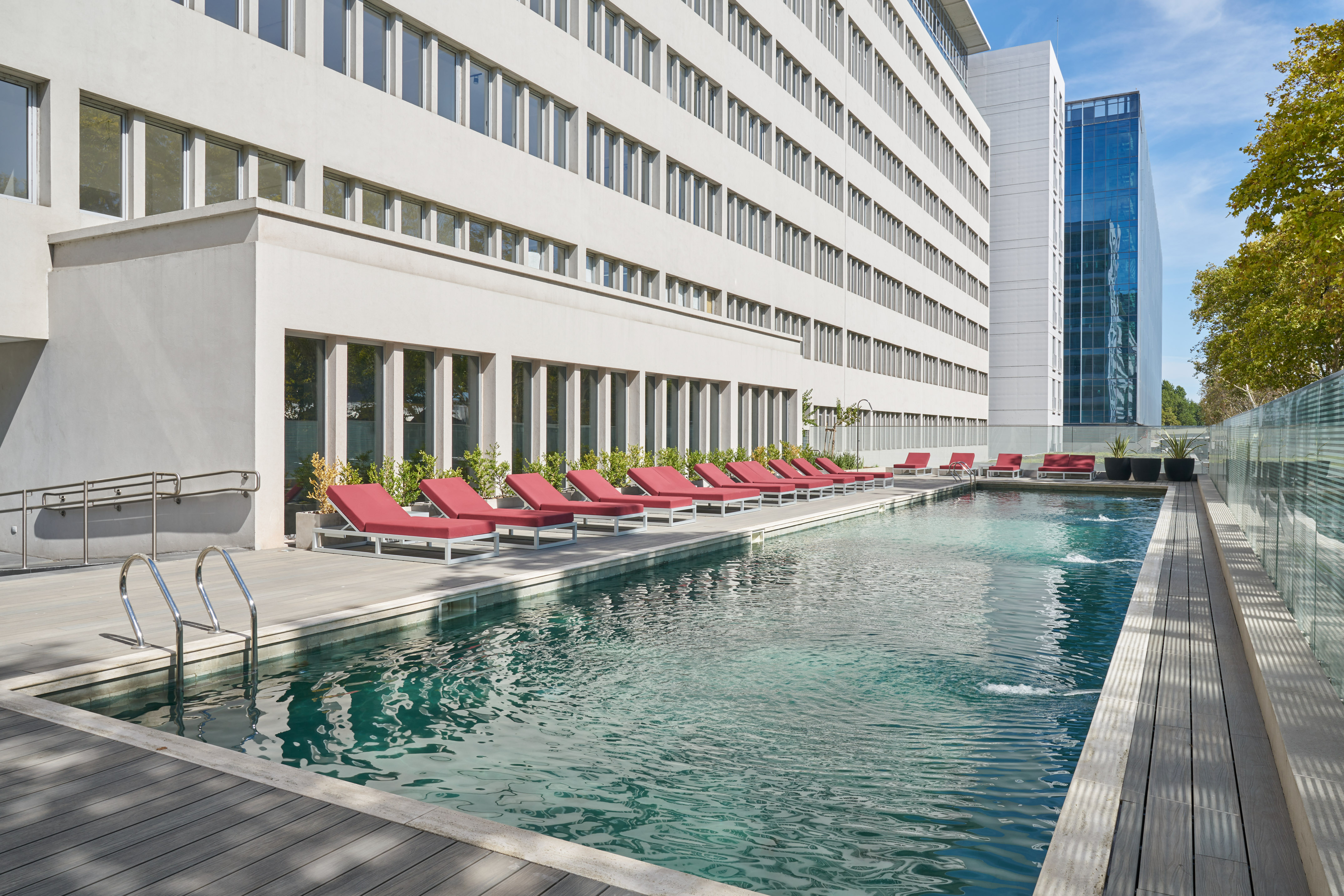 Outdoor pool with red sun loungers on a wooden deck, modern hotel building.