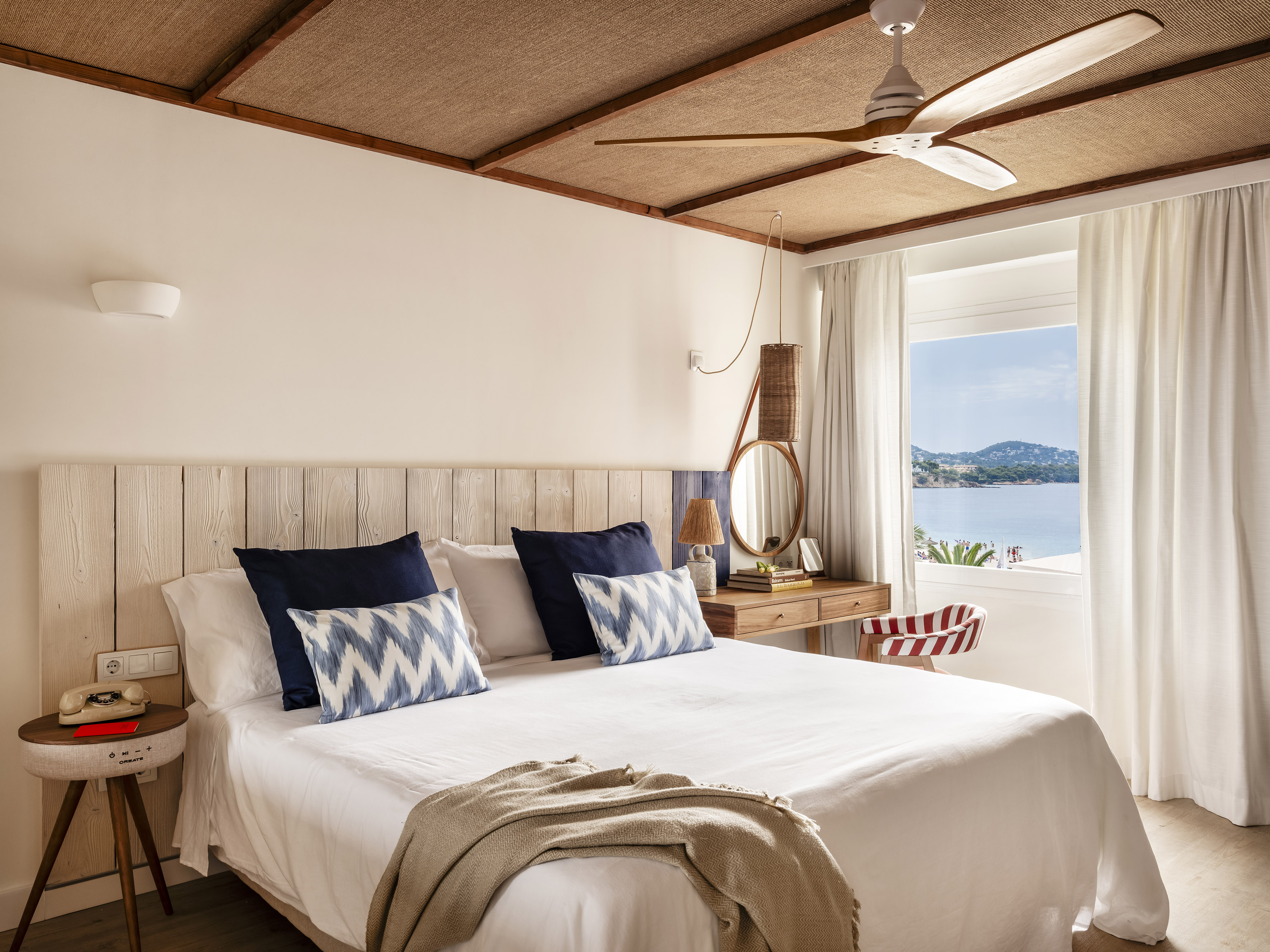 Coastal hotel bedroom with sea view, woven ceiling, wood headboard, and fan.