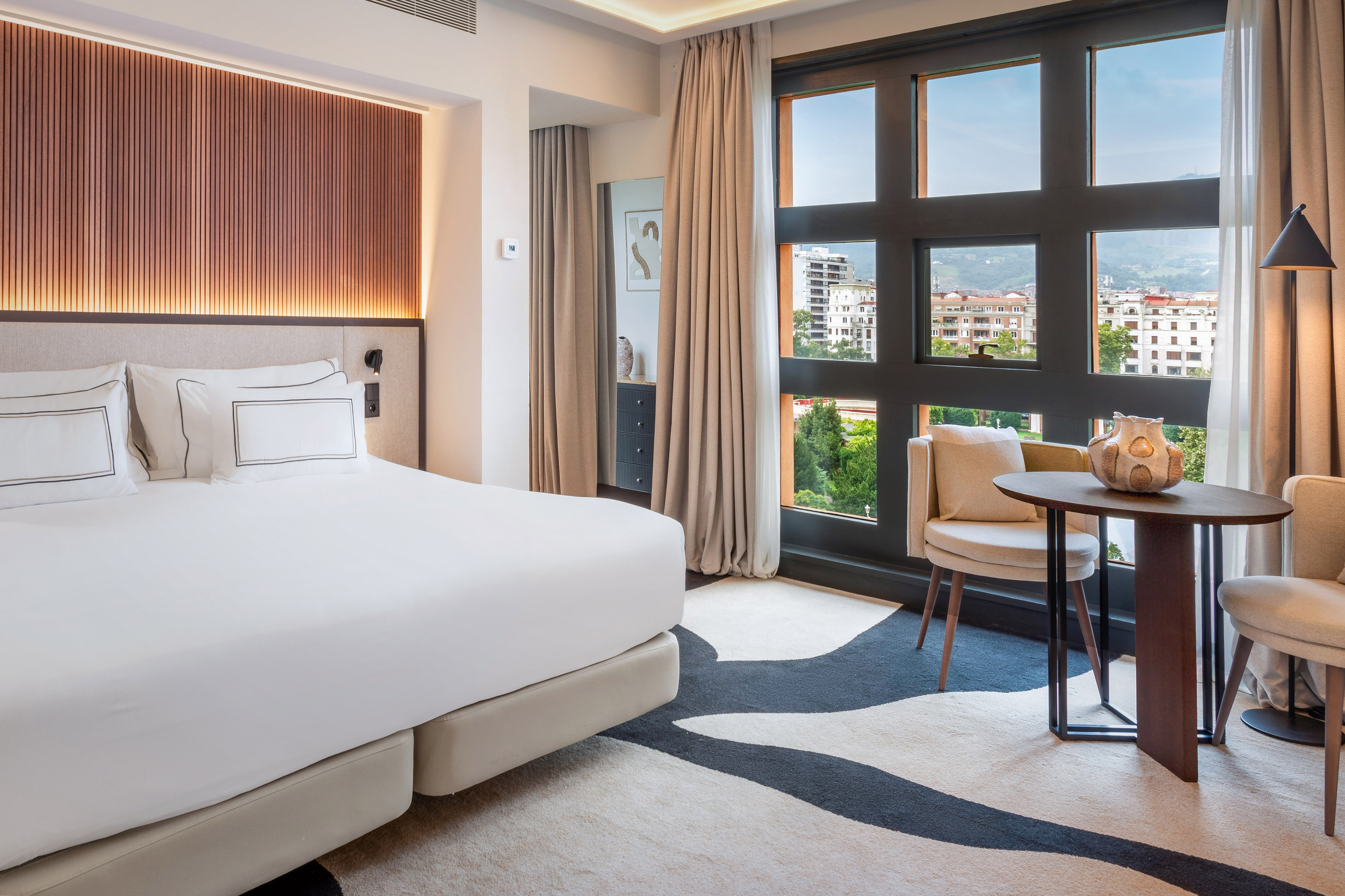 Luxury hotel bedroom with white bed, city view window, and cozy seating area.