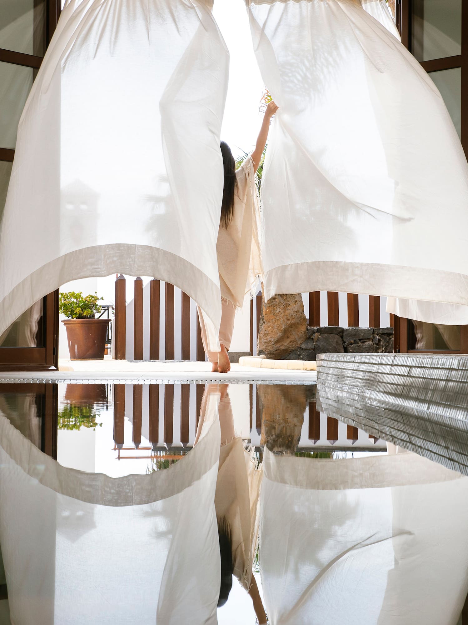 Woman opening sheer white curtains to a bright resort pool with reflections.
