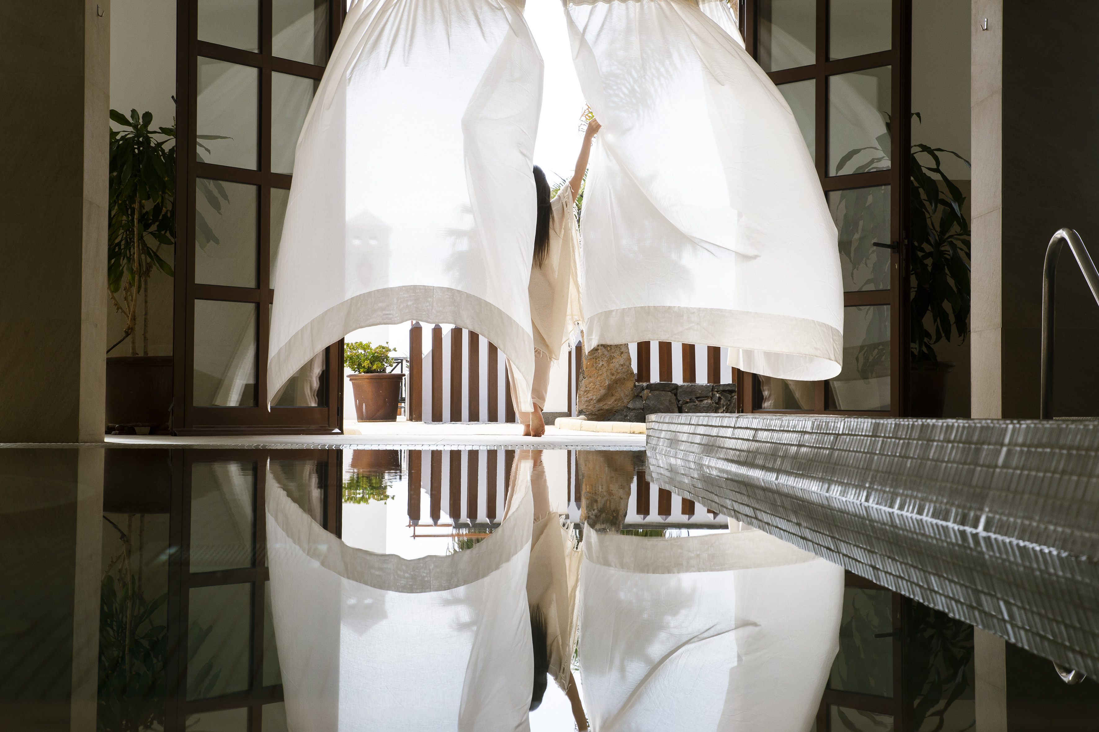 Woman opening sheer white curtains to a bright resort pool with reflections.