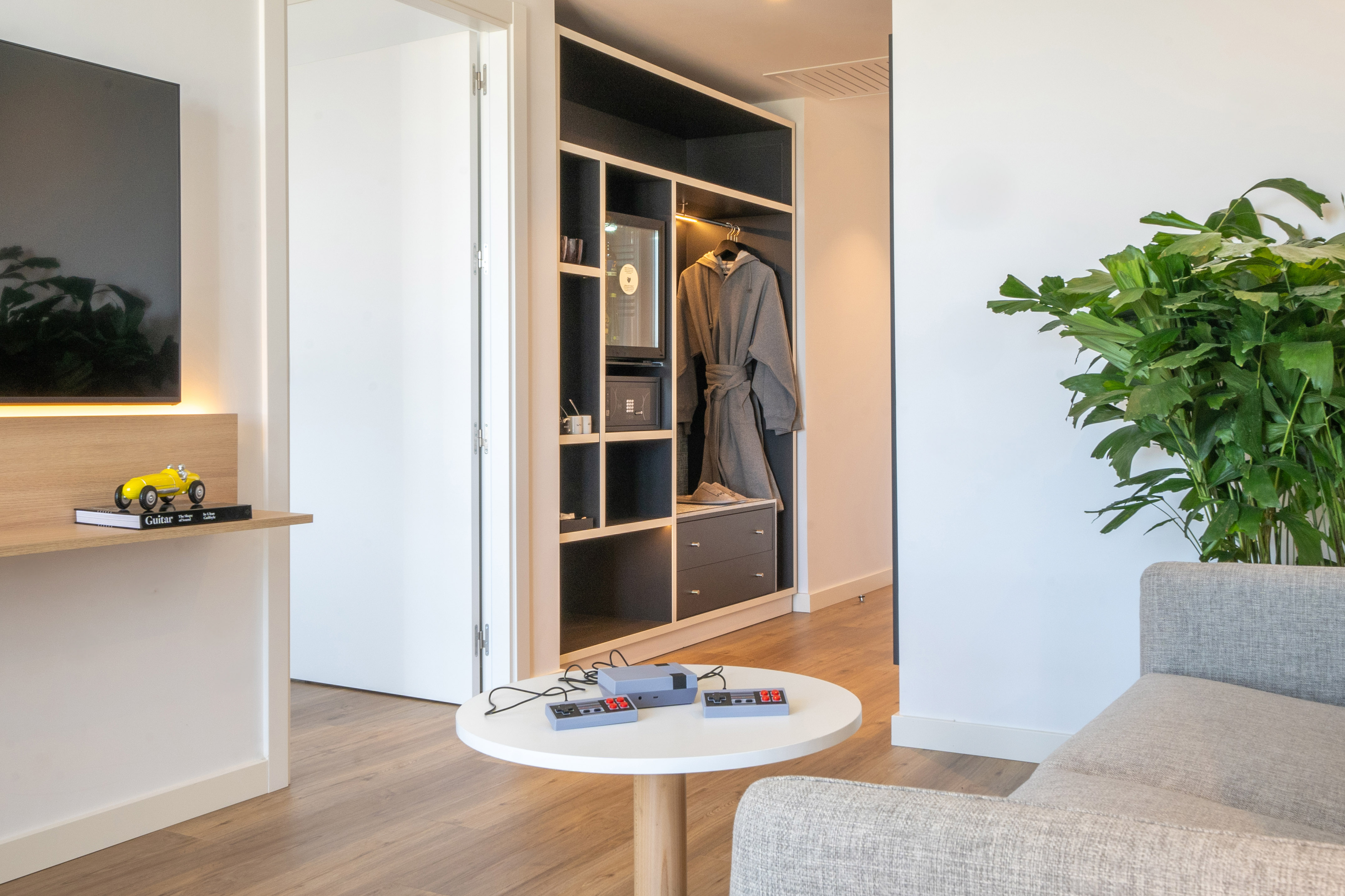 Modern hotel room: living area with retro NES console, gray couch, open wardrobe.