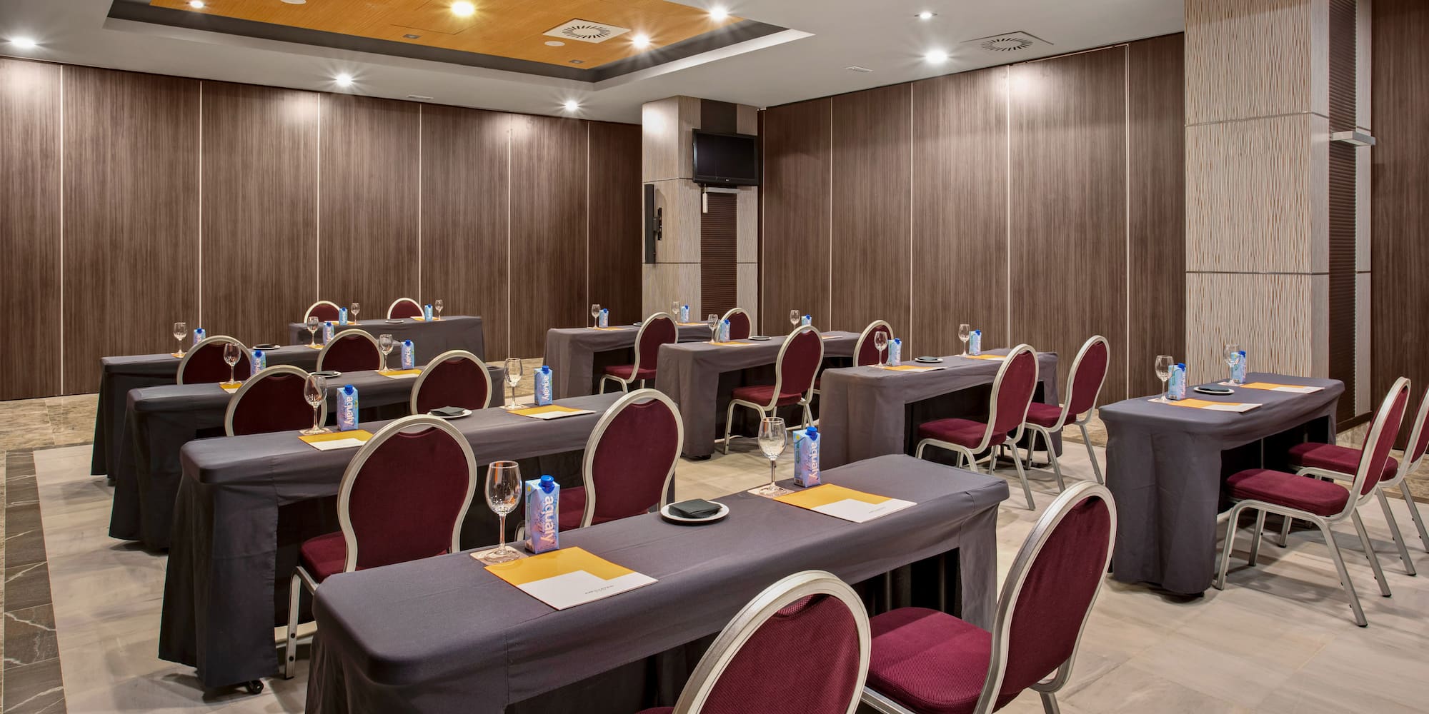 Modern conference room with grey tables, burgundy chairs, wood walls, and TV.