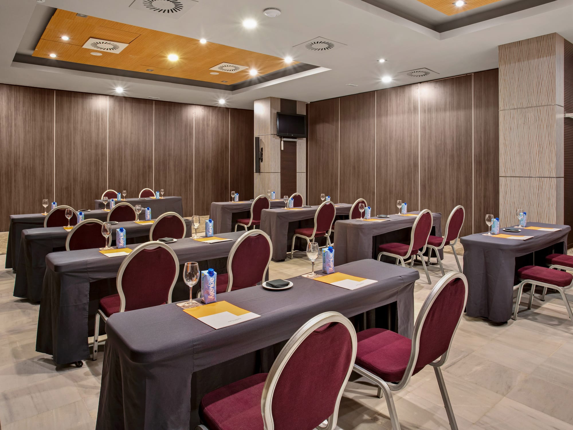 Hotel meeting room with dark tables, burgundy chairs, wood walls, TV screen.