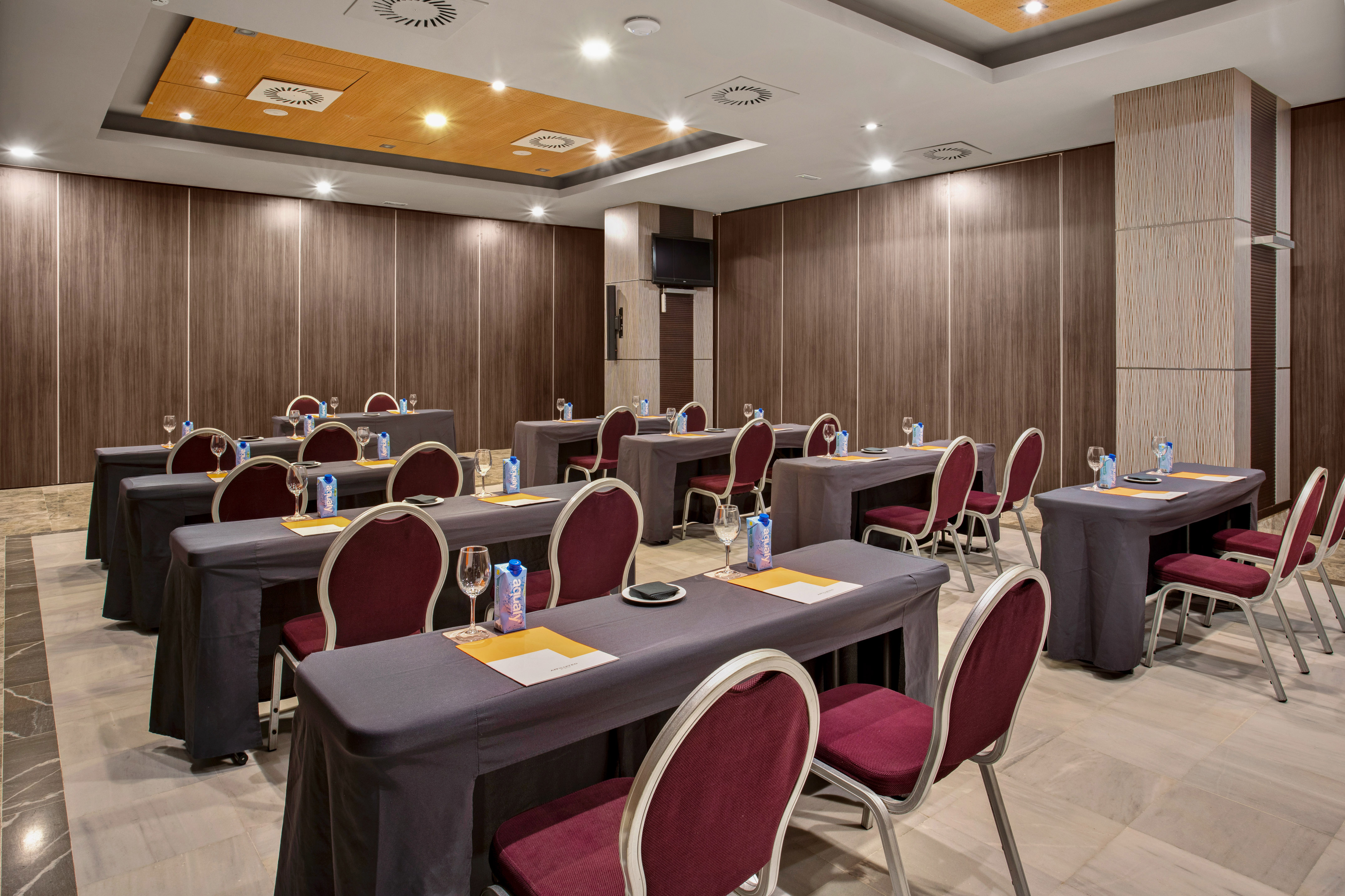 Hotel meeting room with dark tables, burgundy chairs, wood walls, TV screen.