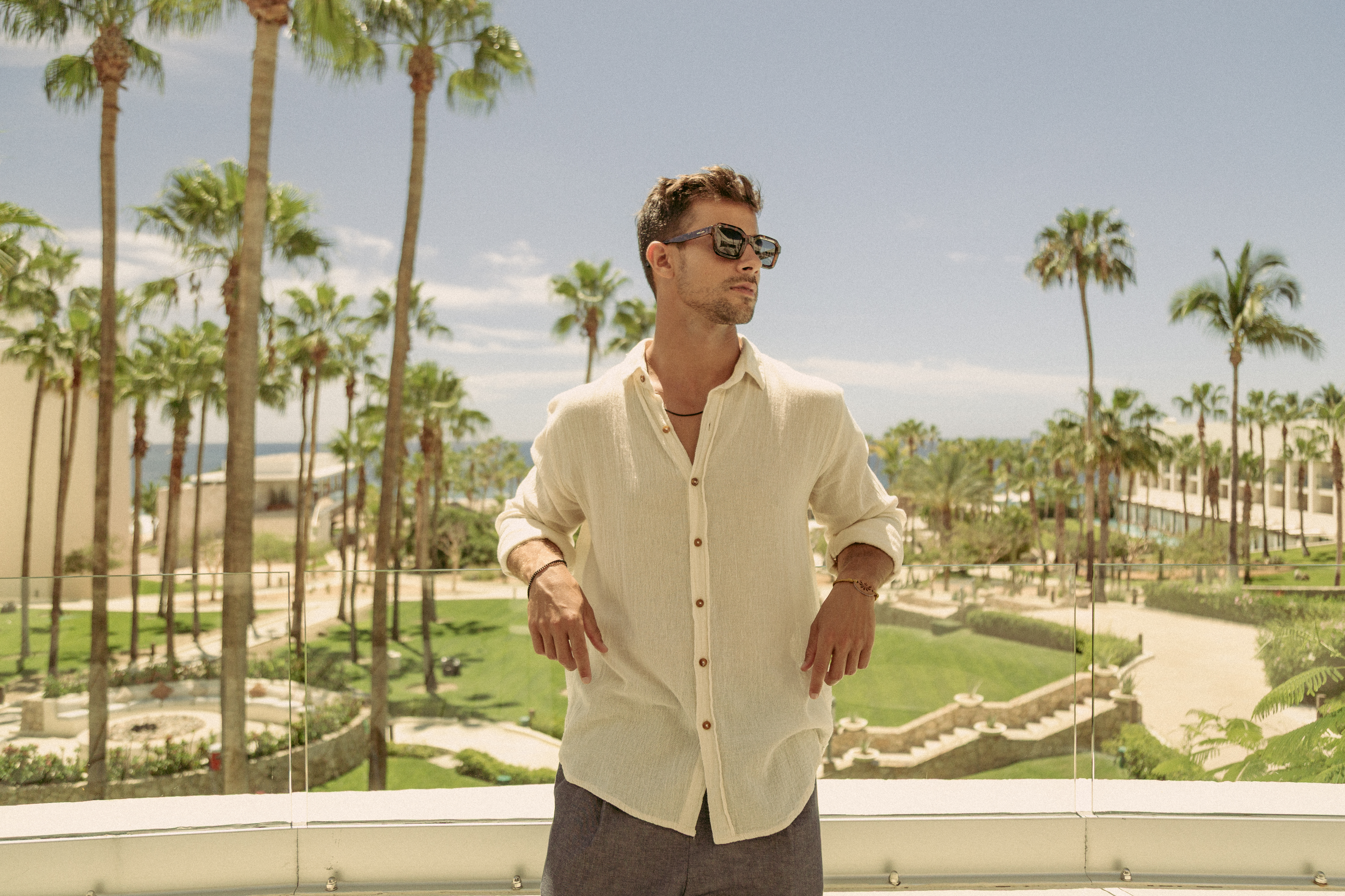 Man in linen shirt, sunglasses on resort balcony with palm trees.