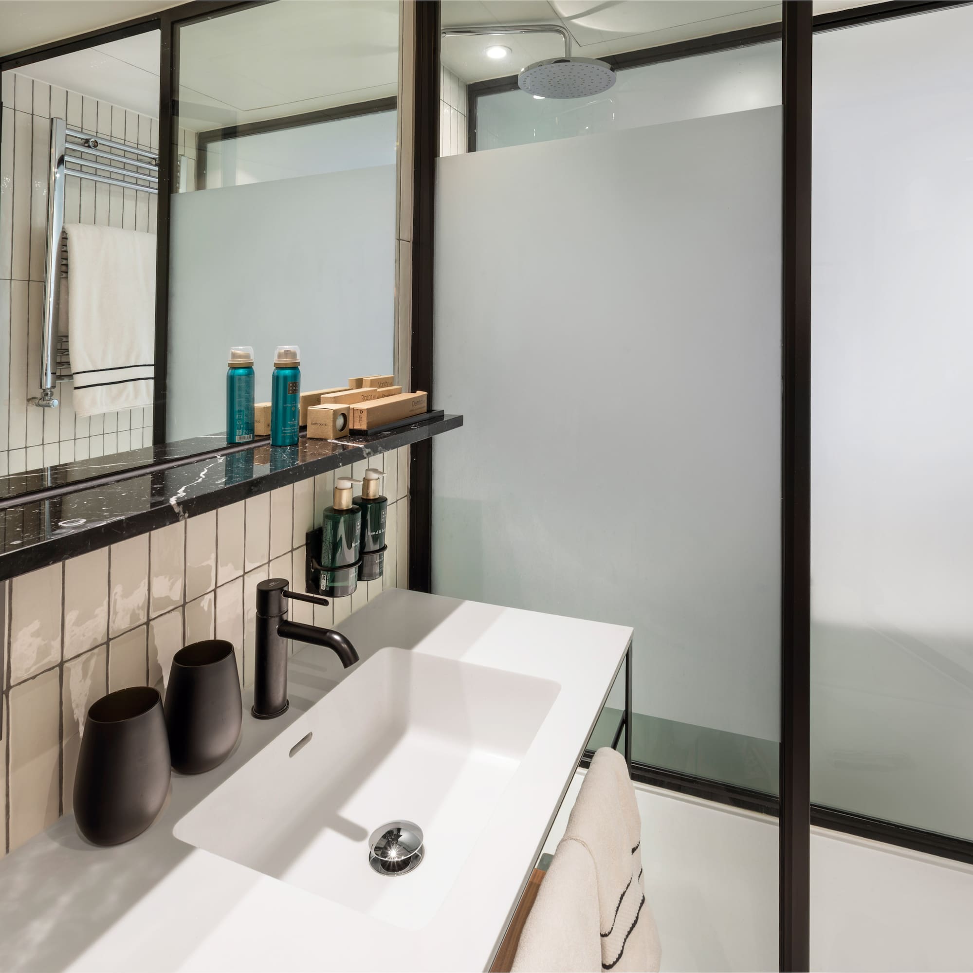 Modern hotel bathroom vanity with white sink, black faucet, and frosted glass shower.