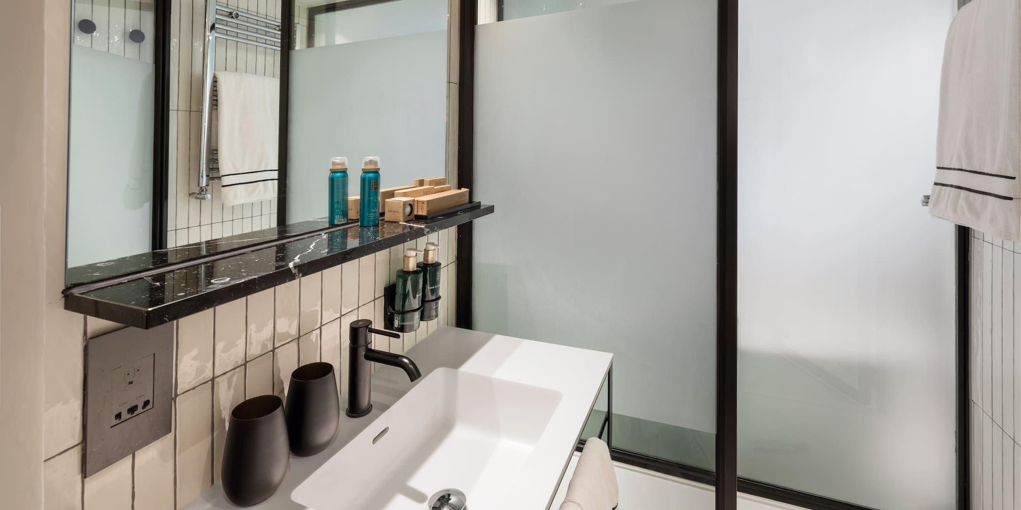 Modern hotel bathroom vanity with white sink, black faucet, and frosted glass shower.