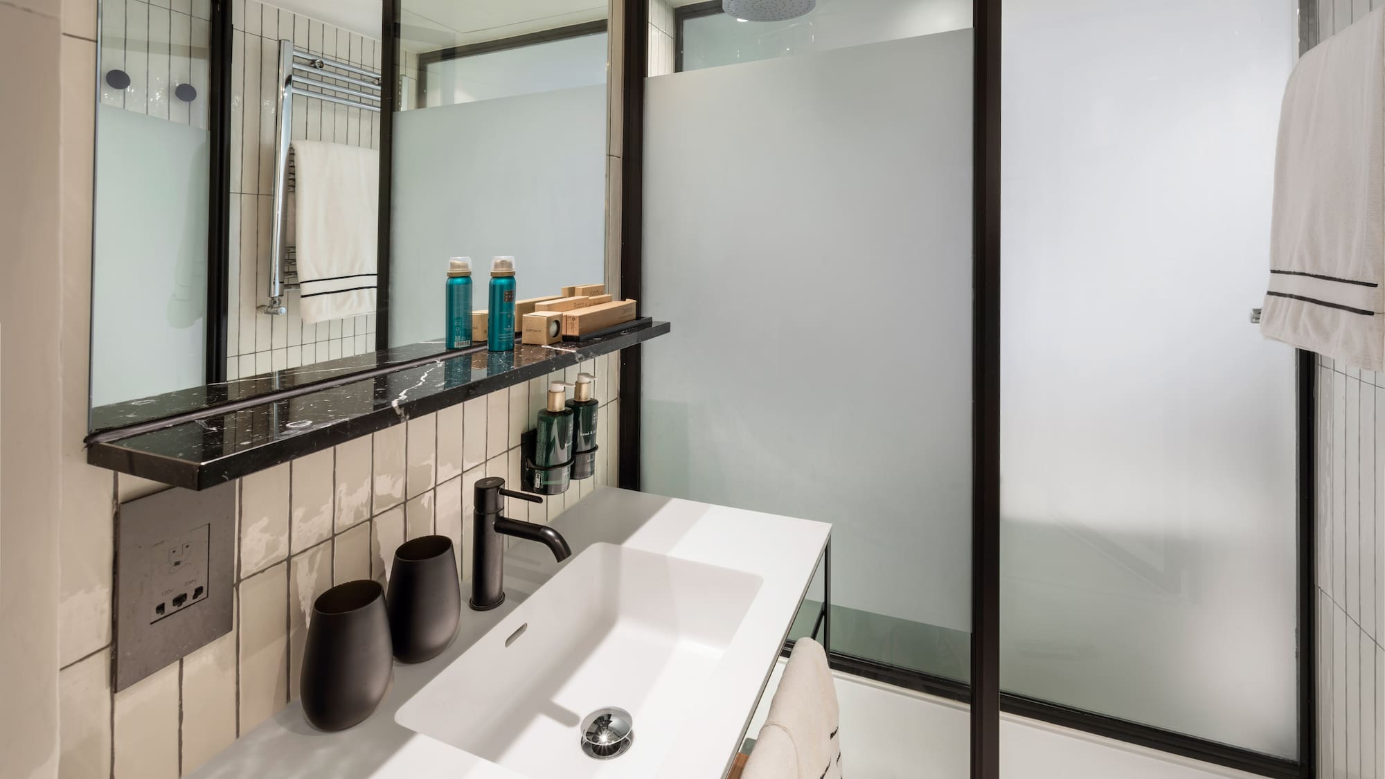 Modern hotel bathroom vanity with white sink, black faucet, and frosted glass shower.