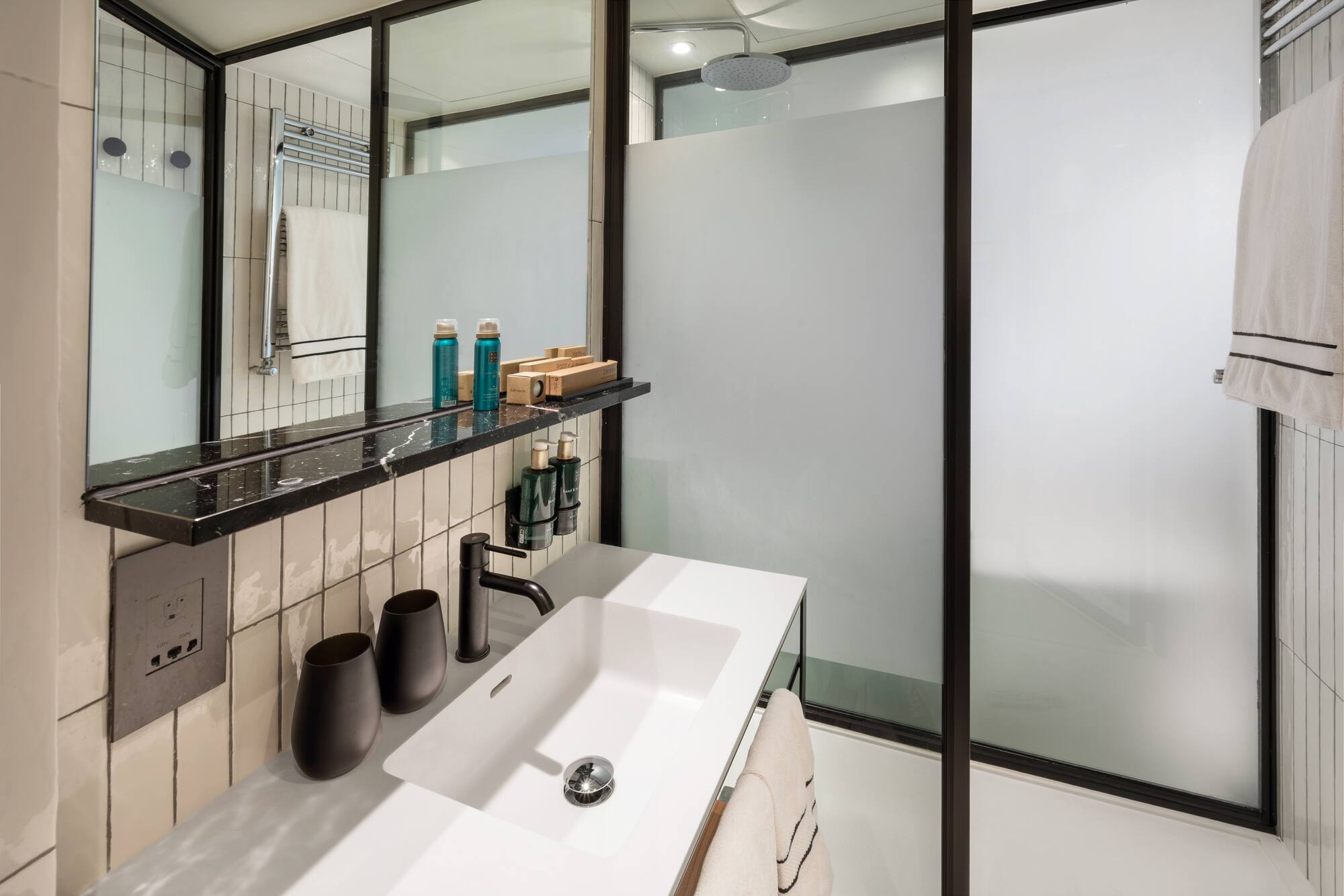 Modern hotel bathroom vanity with white sink, black faucet, and frosted glass shower.