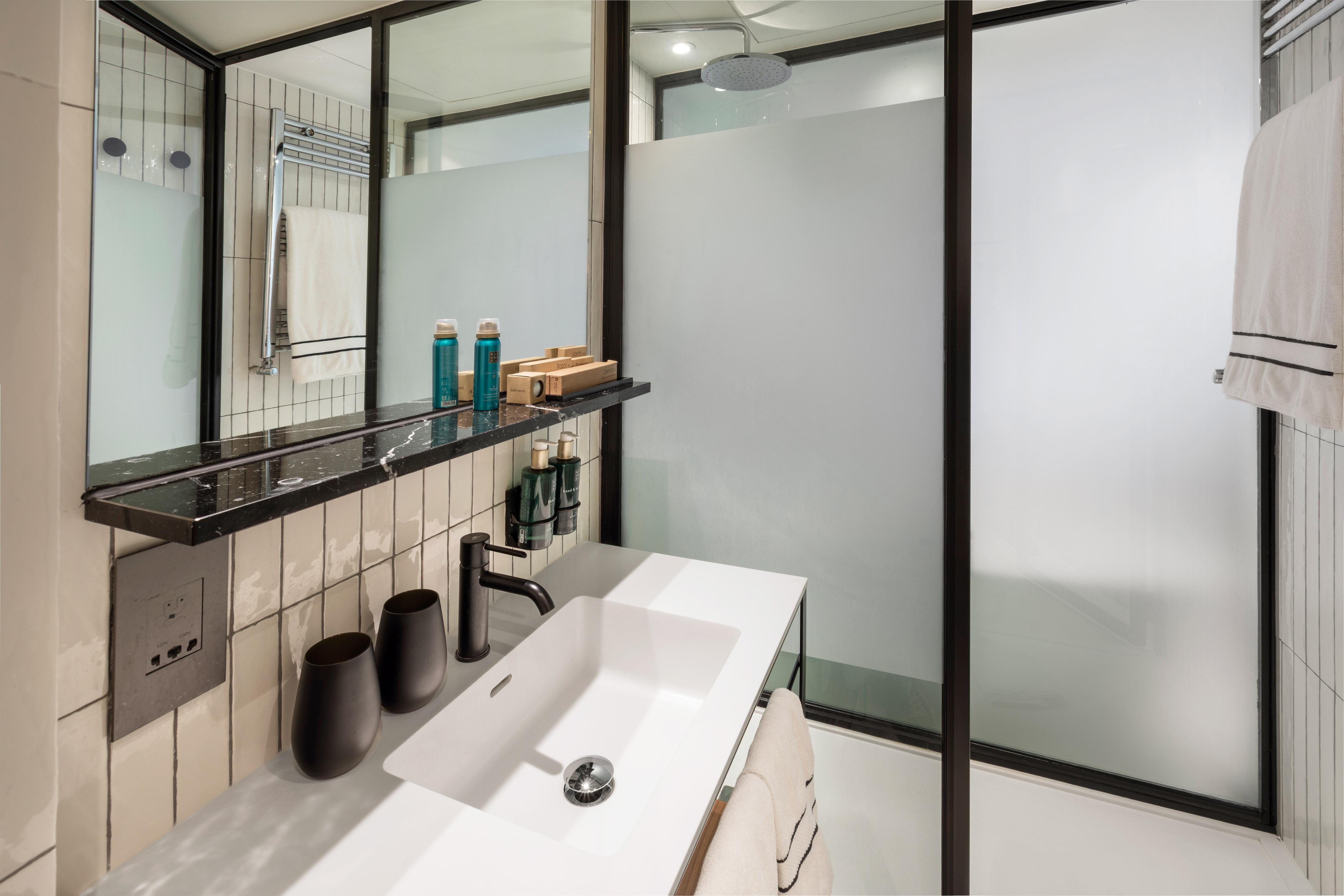 Modern hotel bathroom vanity with white sink, black faucet, and frosted glass shower.