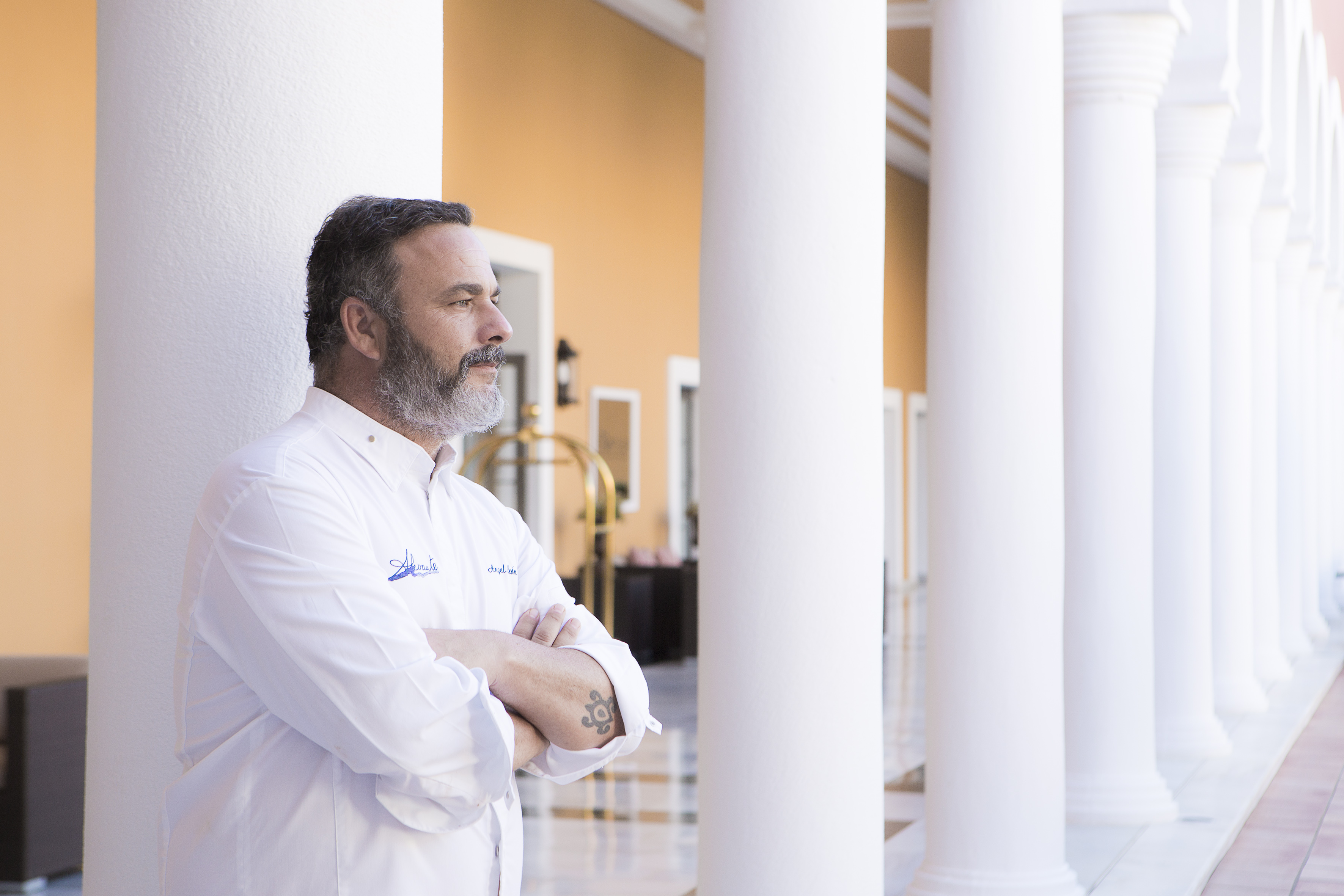 Bearded chef in white uniform standing by hotel colonnade.