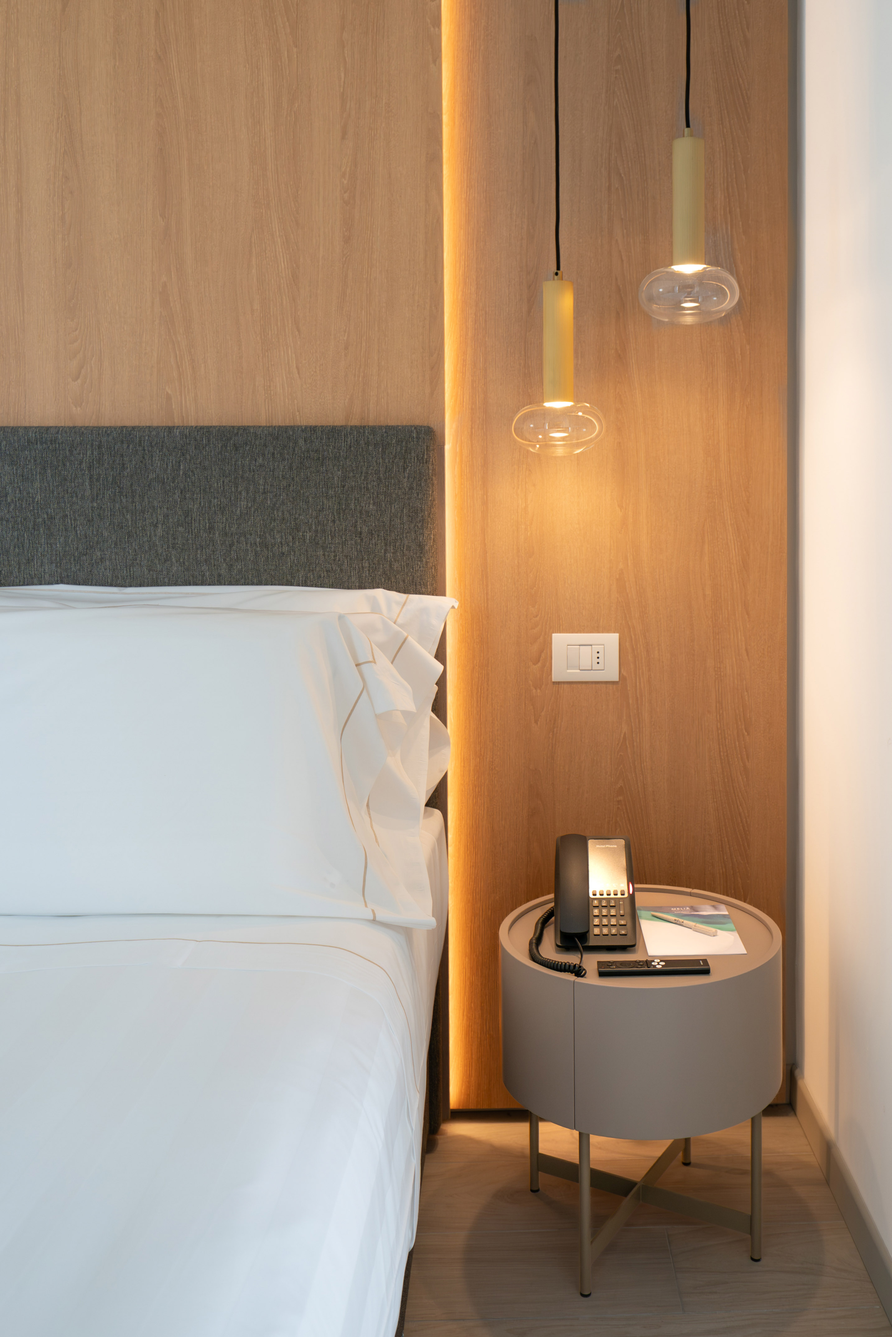 Modern hotel bedroom: white bed, wood paneling, pendant lights, nightstand.
