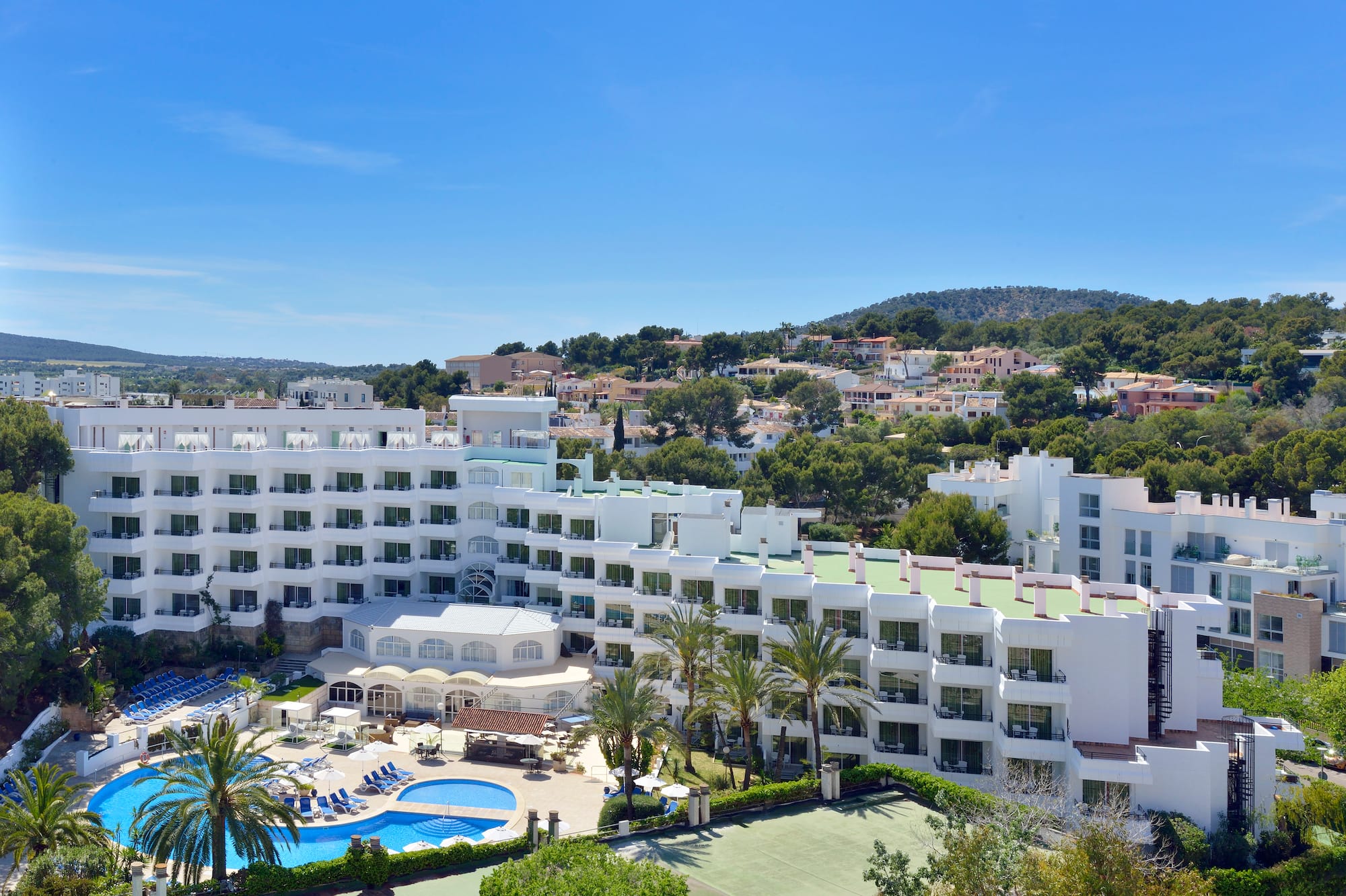 Sunny resort hotel with large swimming pool, palm trees, and tennis court.