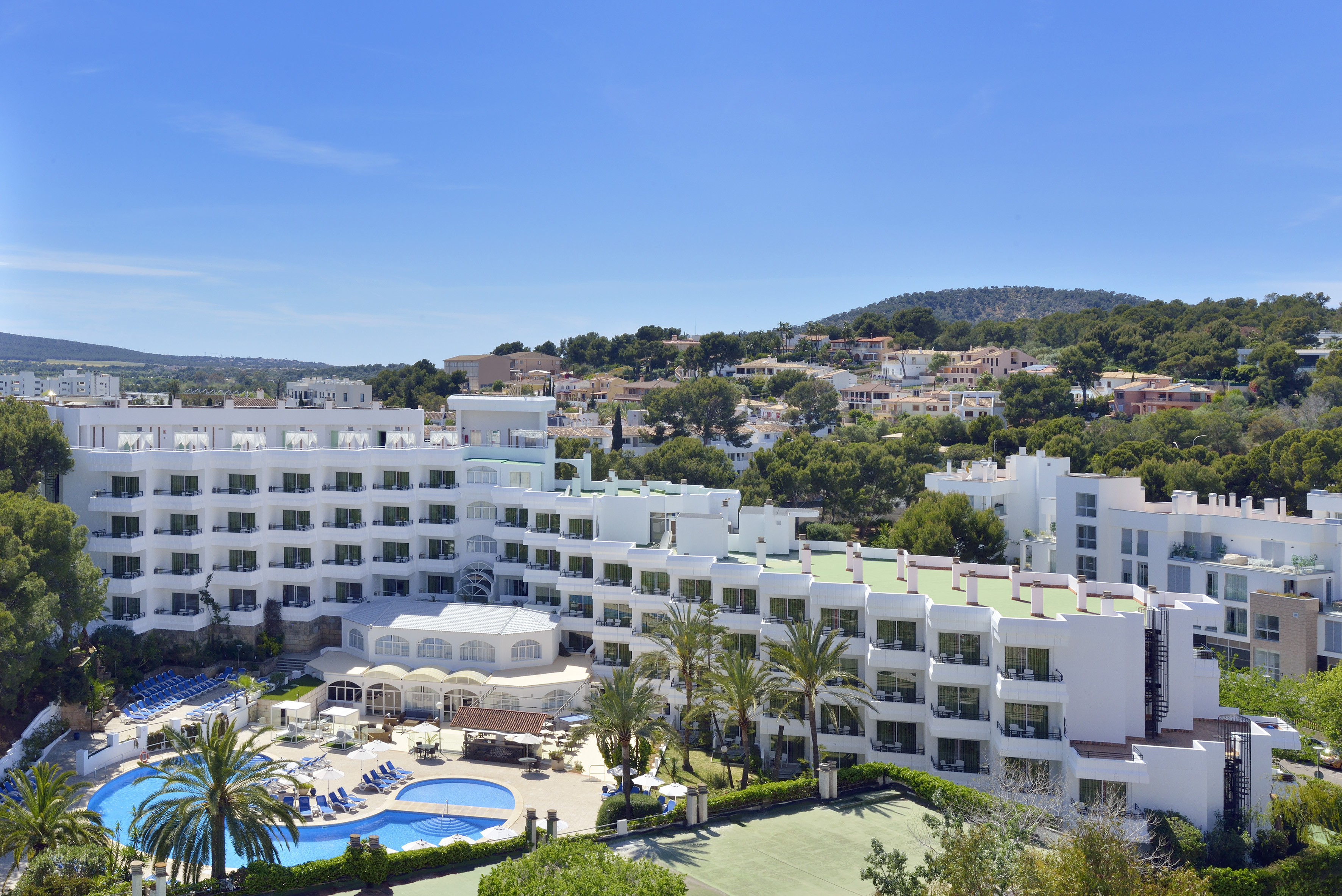 Sunny resort hotel with large swimming pool, palm trees, and tennis court.