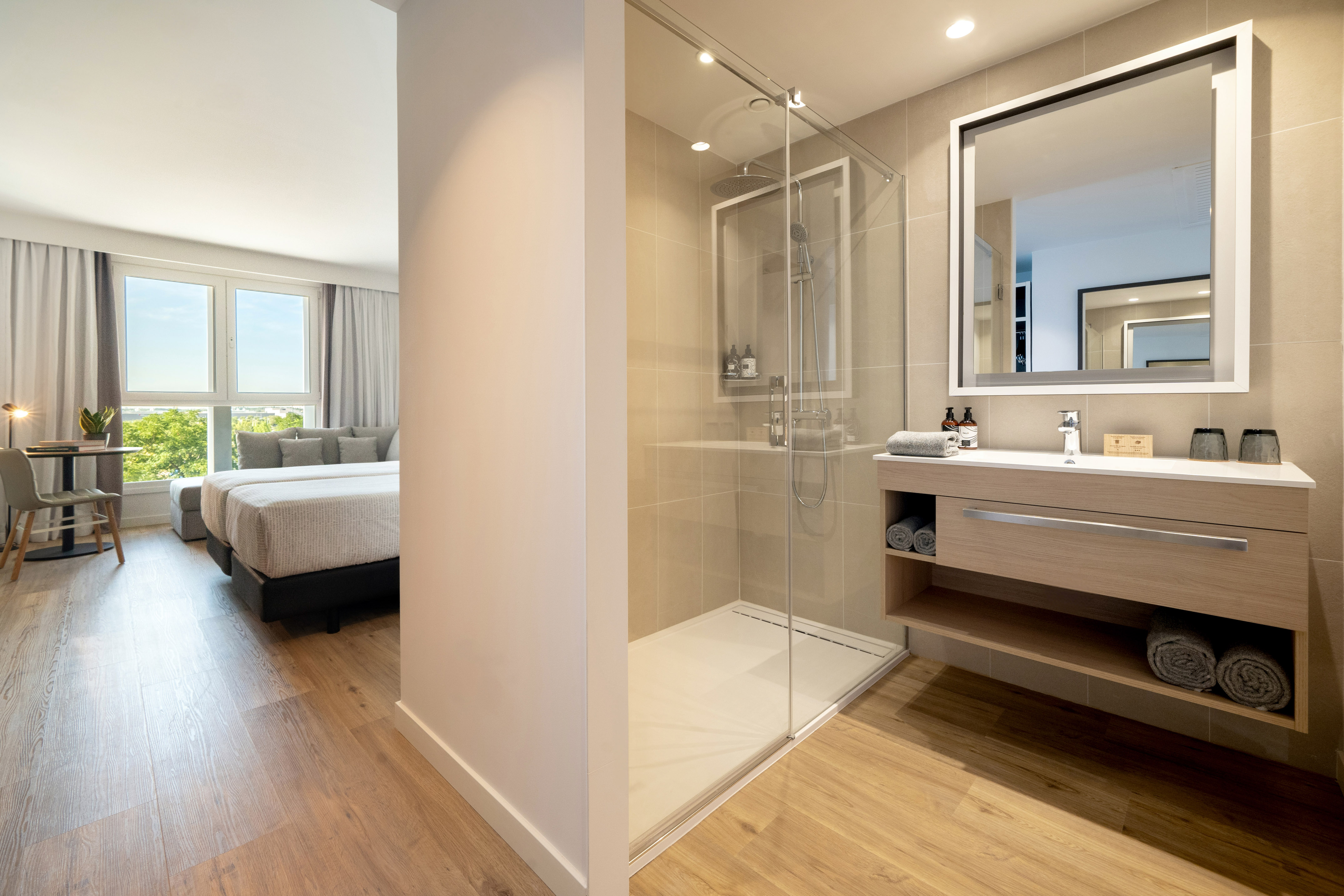 Modern hotel suite with glass shower, vanity, bed, and bright window.