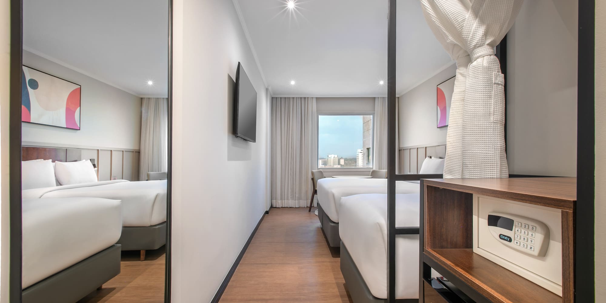 Modern hotel room with twin beds, city view, wall TV, bathrobe, and safe.