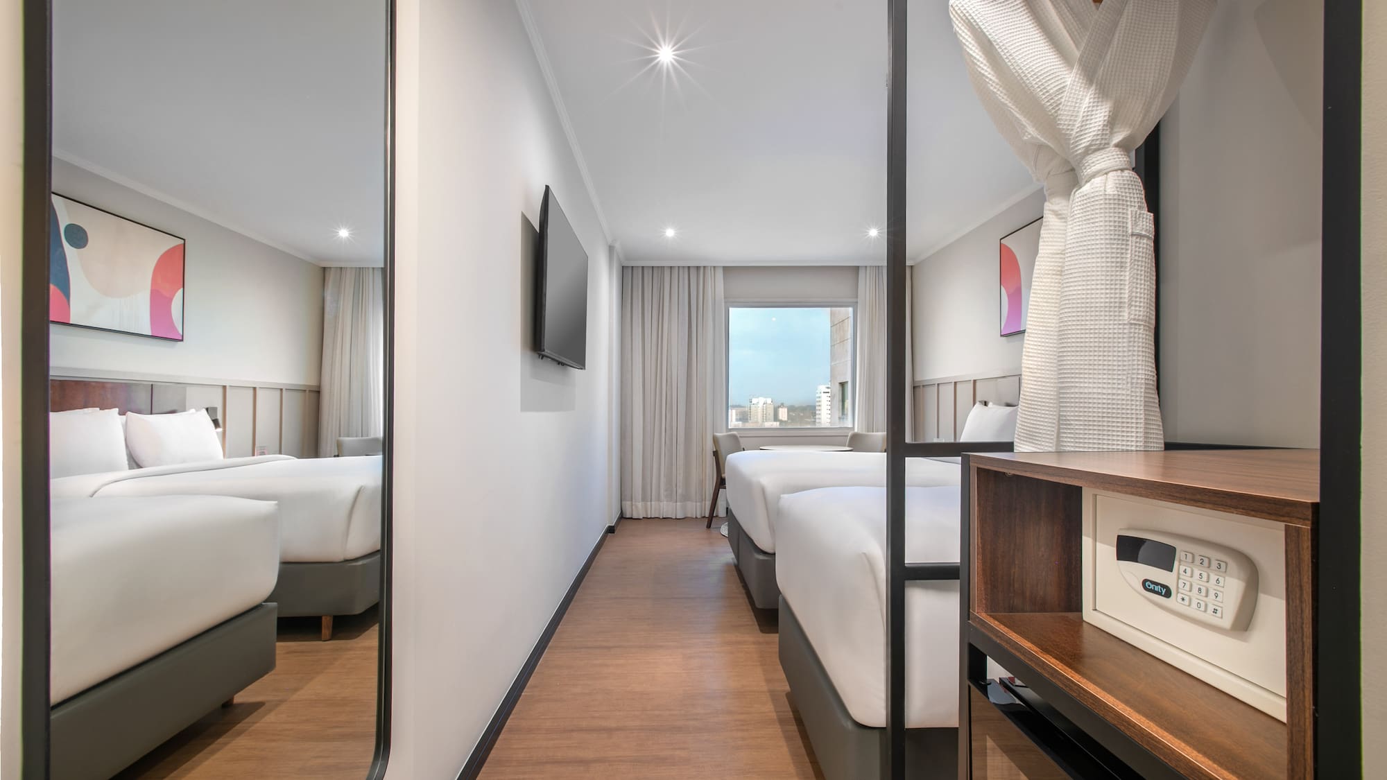 Modern hotel room with twin beds, city view, wall TV, bathrobe, and safe.