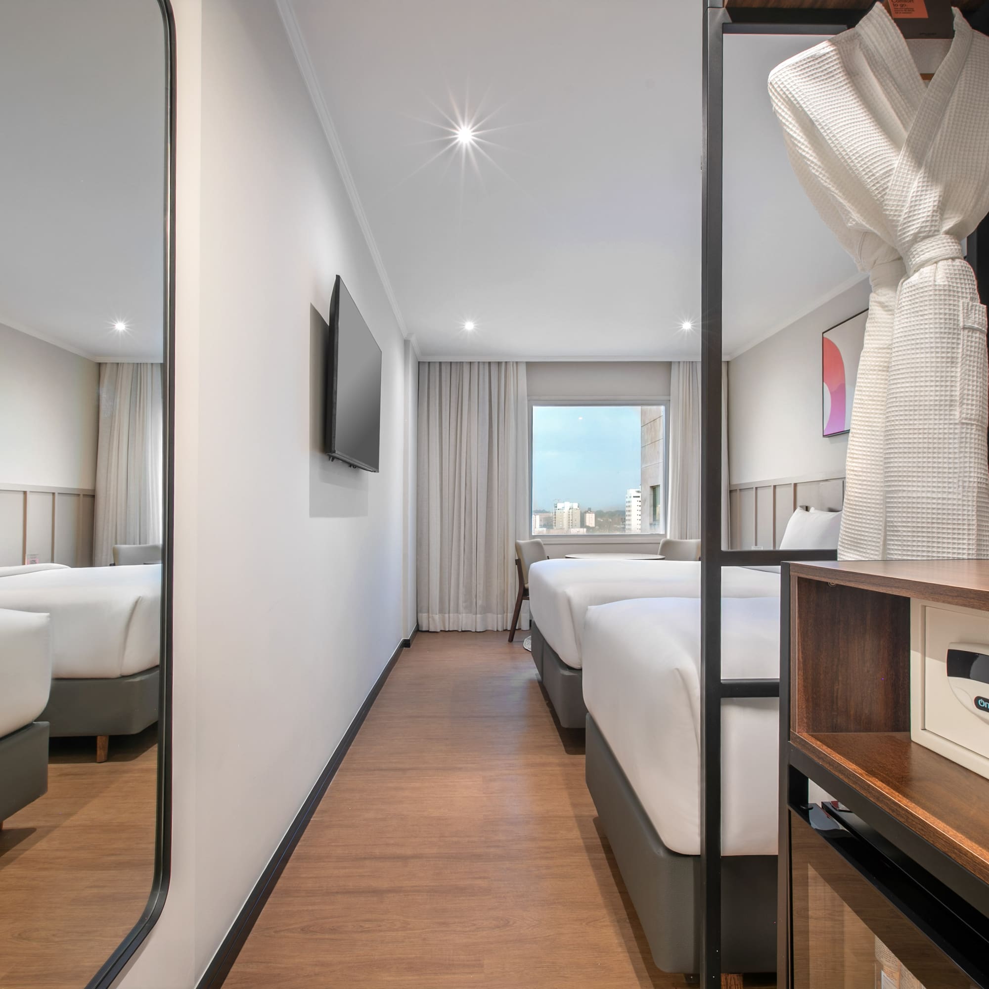 Modern hotel room with twin beds, city view, wall TV, bathrobe, and safe.