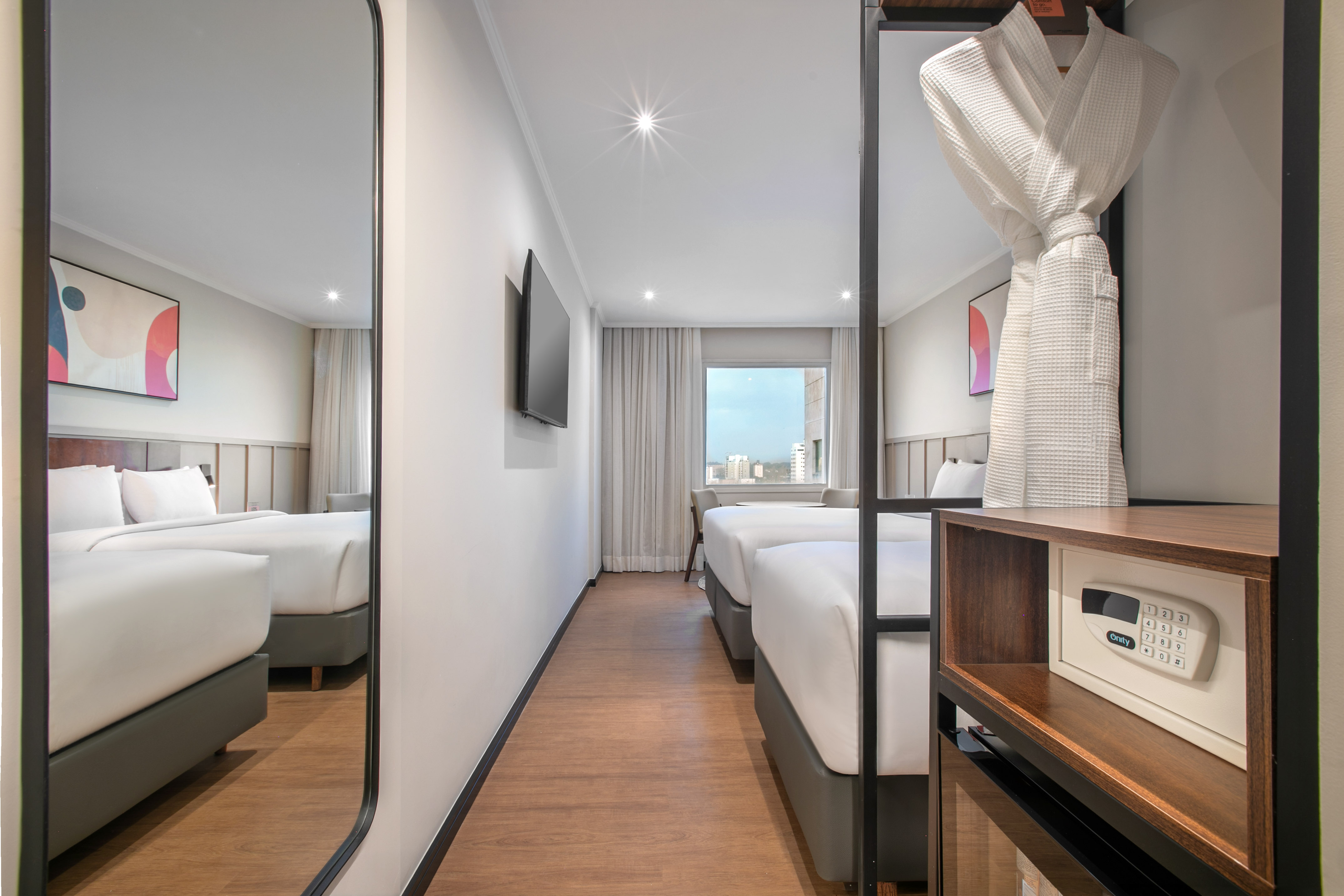 Modern hotel room with twin beds, city view, wall TV, bathrobe, and safe.