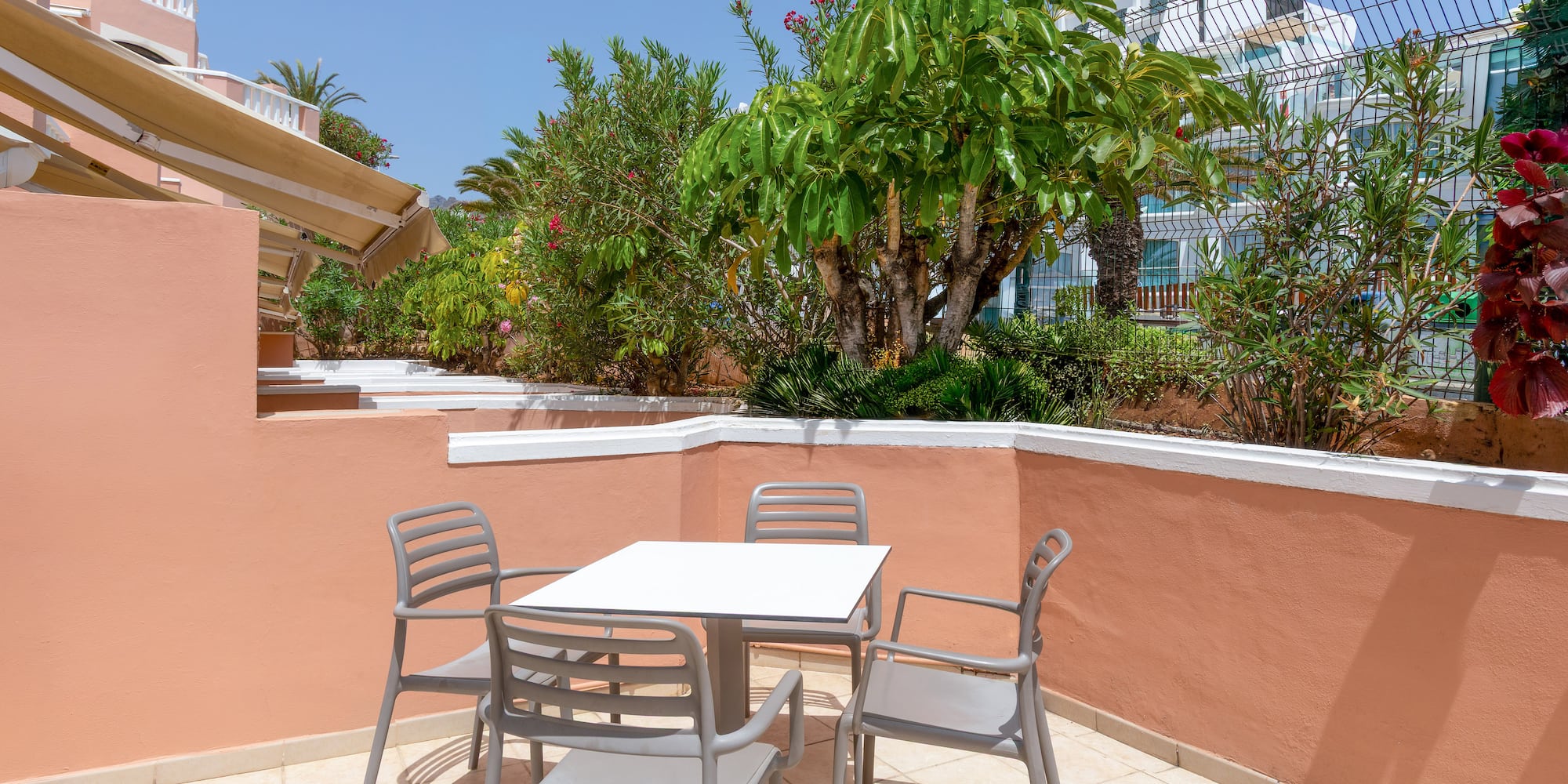 Sunny hotel patio with outdoor dining table, chairs, and lush tropical plants.