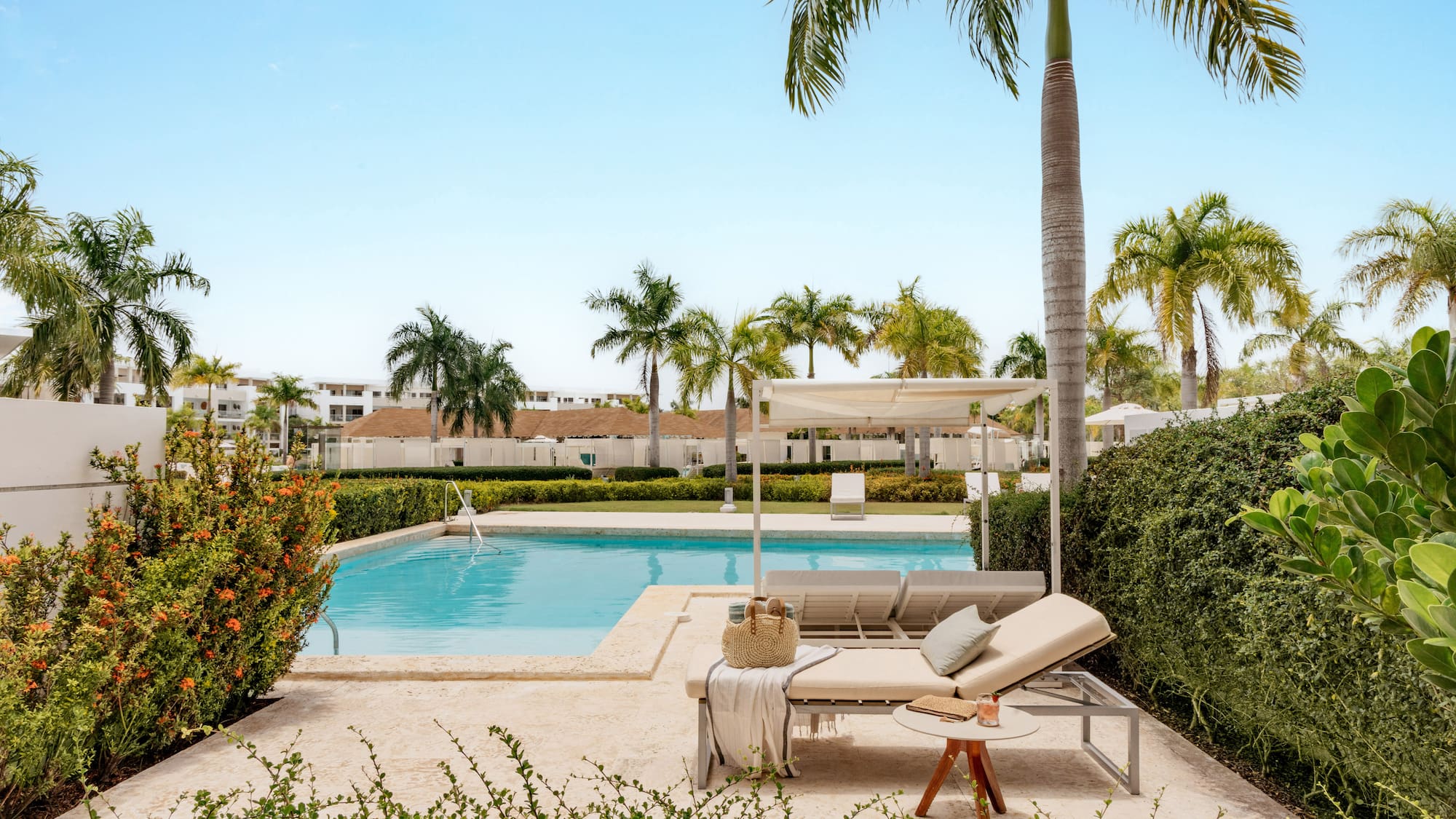 Tropical resort pool, daybeds, palm trees, and lush green landscaping.