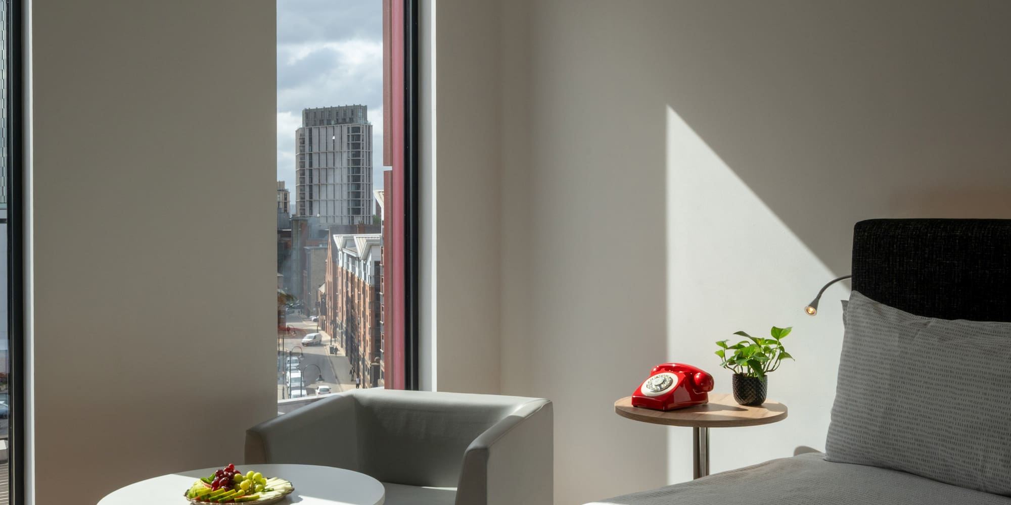 Modern hotel bedroom with city view, fruit plate, and red rotary phone.