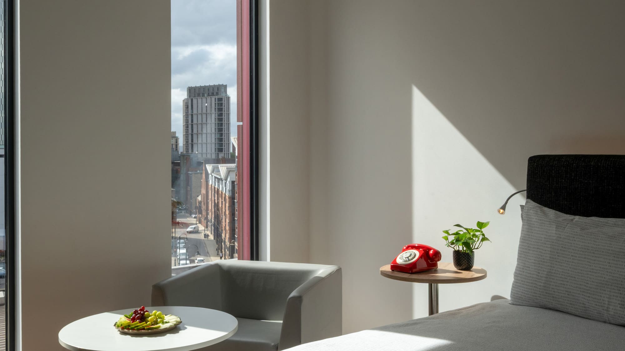 Modern hotel bedroom with city view, fruit plate, and red rotary phone.