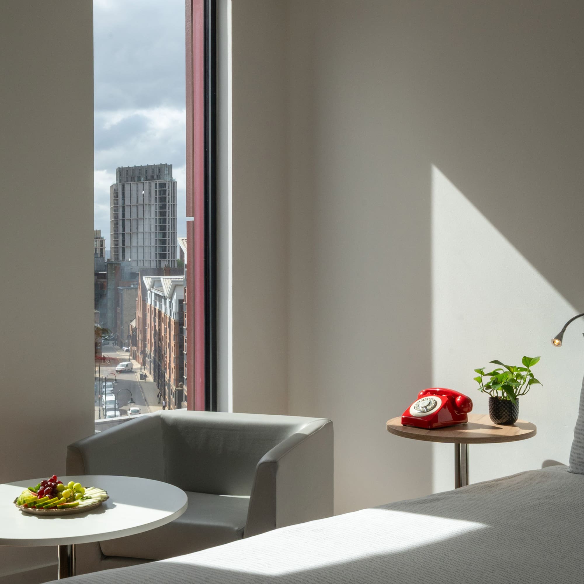 Modern hotel bedroom with city view, fruit plate, and red rotary phone.